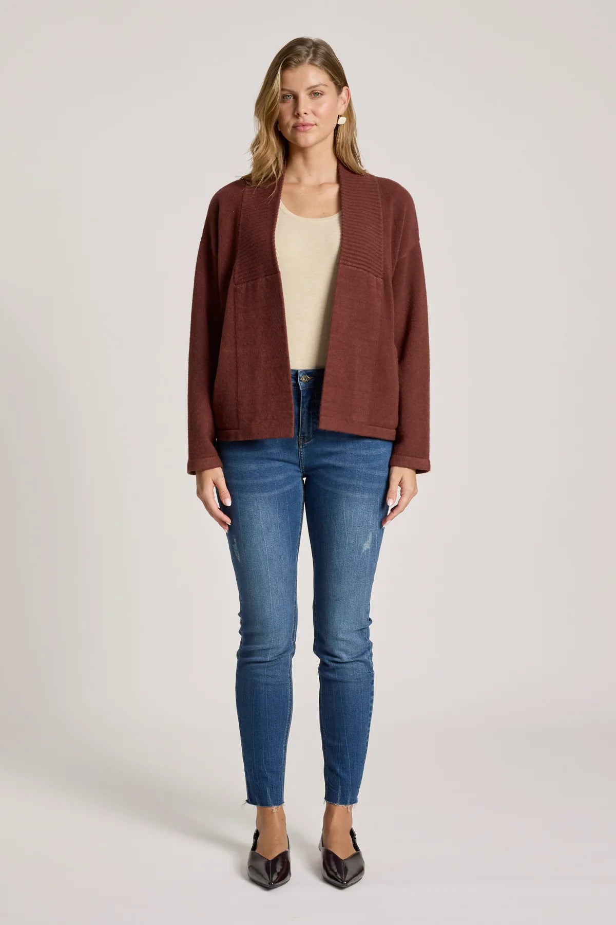 Woman wearing a Eb & Ive brown jacket, beige top, and blue jeans on a white background
