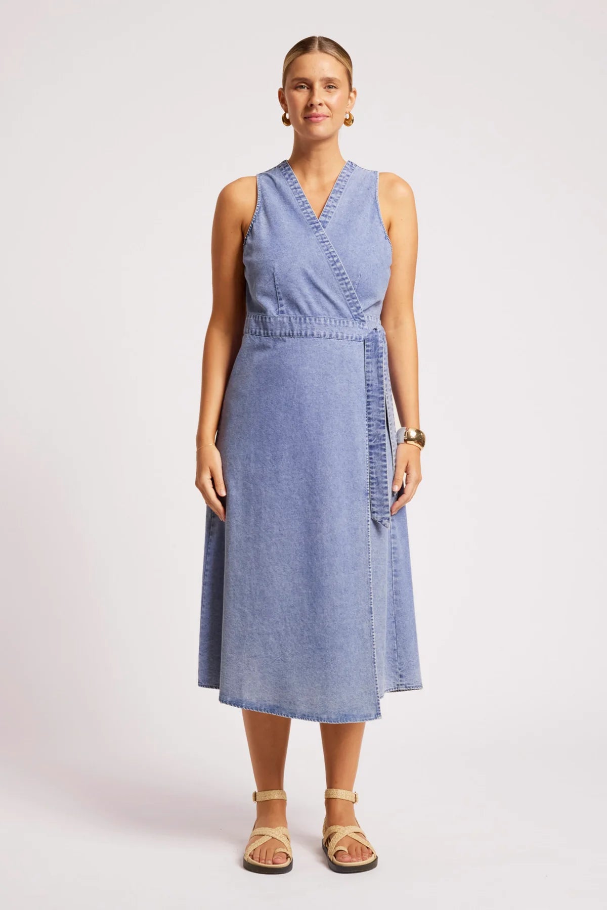 A woman modeling a Eb & Ive sleeveless light blue denim midi wrap dress with a V-neckline and side tie detail, paired with beige strappy sandals against a light gray studio background.