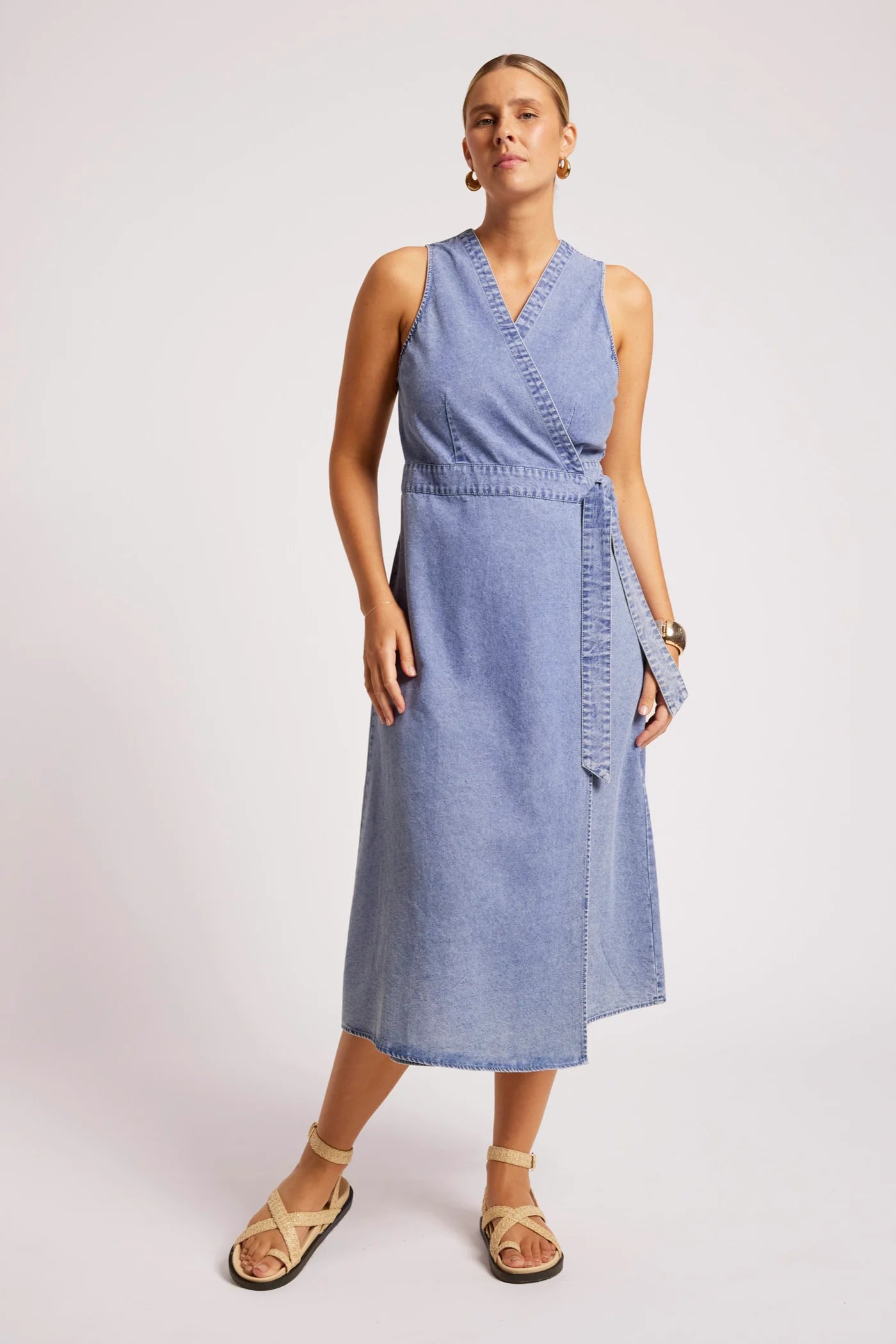 Image of A woman modeling a Eb & Ive sleeveless light blue denim midi wrap dress with a tie waist, paired with beige strappy sandals, against a light gray studio background.