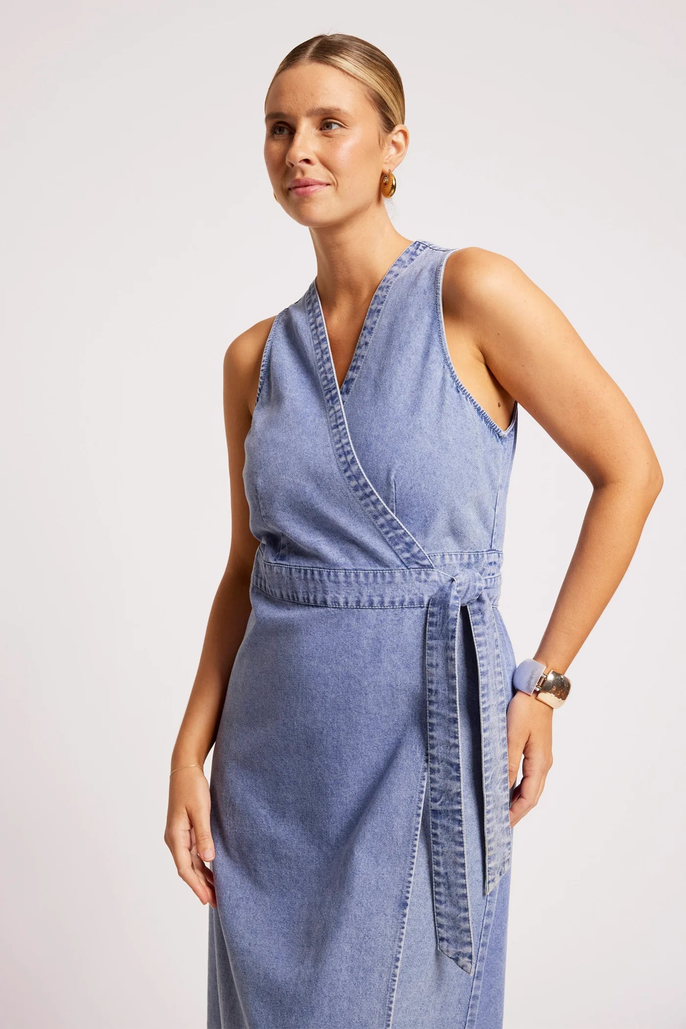 Image of A woman modeling a Eb & Ive sleeveless wrap-style denim dress in a dusty blue shade, featuring a matching tie belt at the waist, set against a plain light gray studio background.