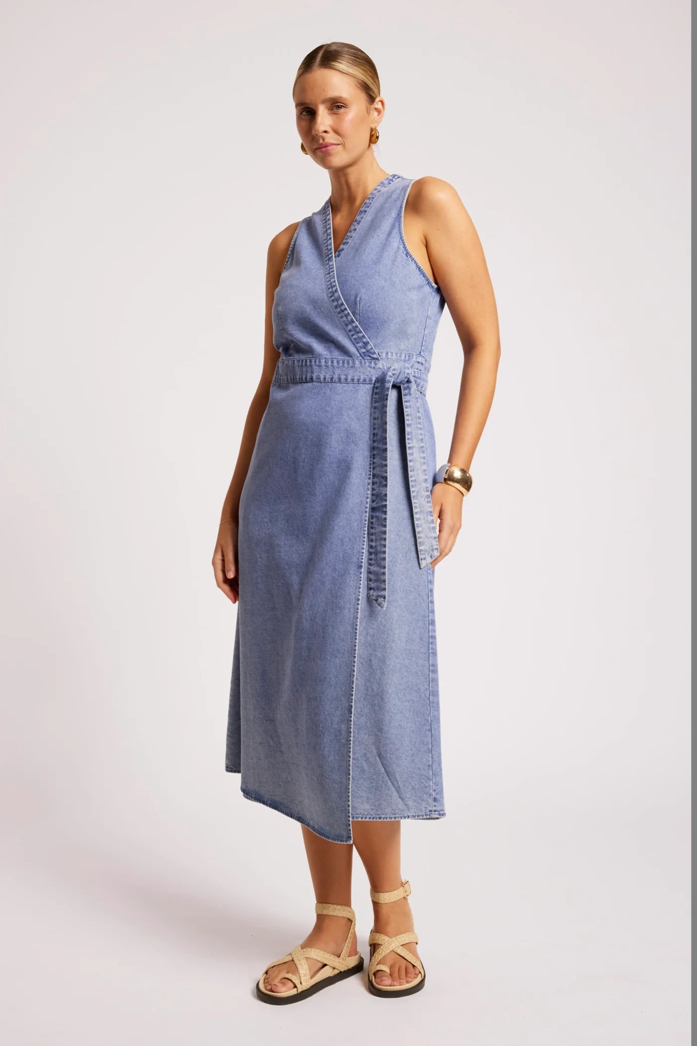 Image of A woman modeling a Eb & Ive sleeveless light blue denim midi dress with a wrap-style V-neck, side tie belt, and side pockets, paired with beige strappy sandals against a plain light gray studio background.