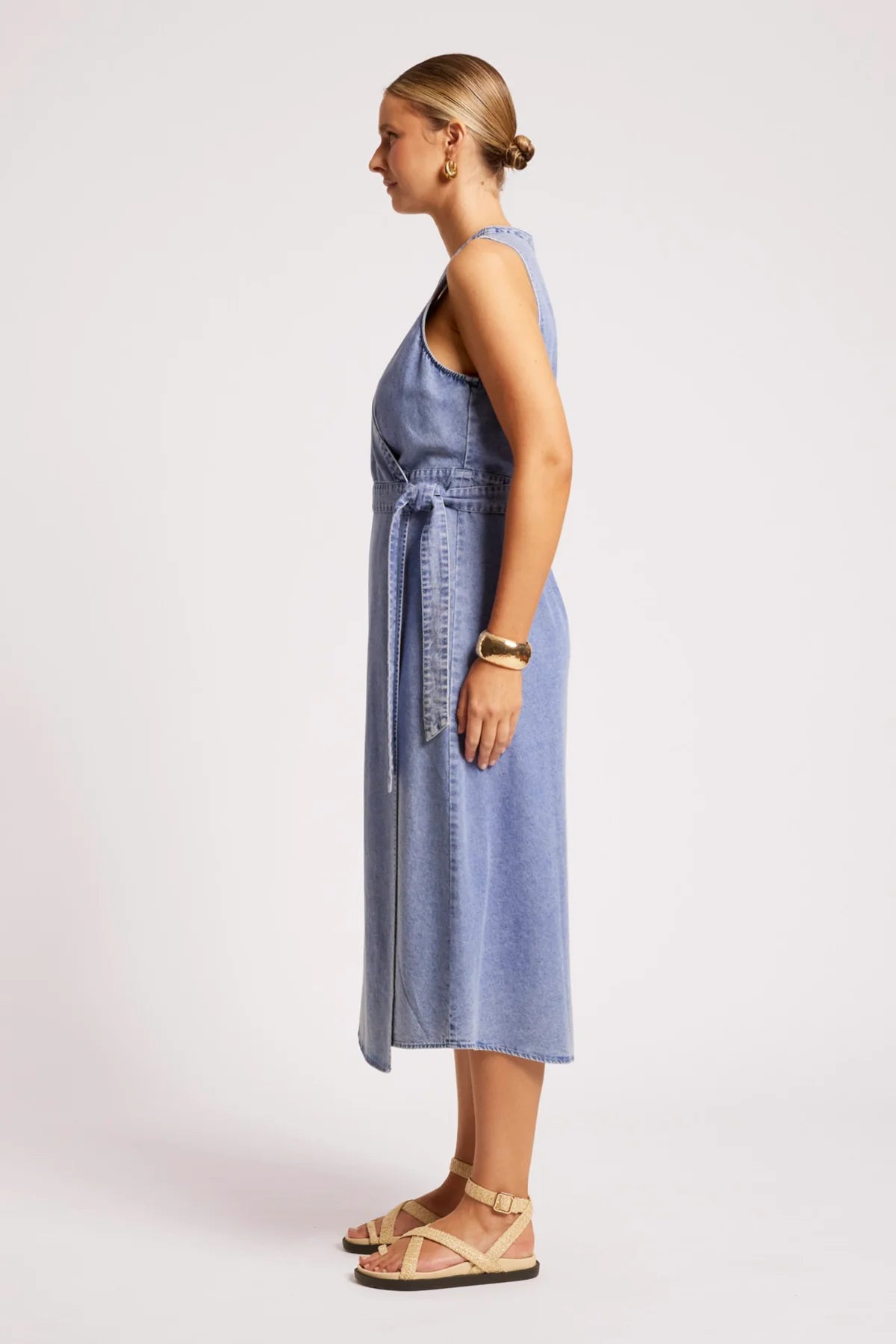 A woman modeling a Eb & Ive sleeveless light blue denim midi dress with a tie waist, shown in profile against a plain light gray studio background.
