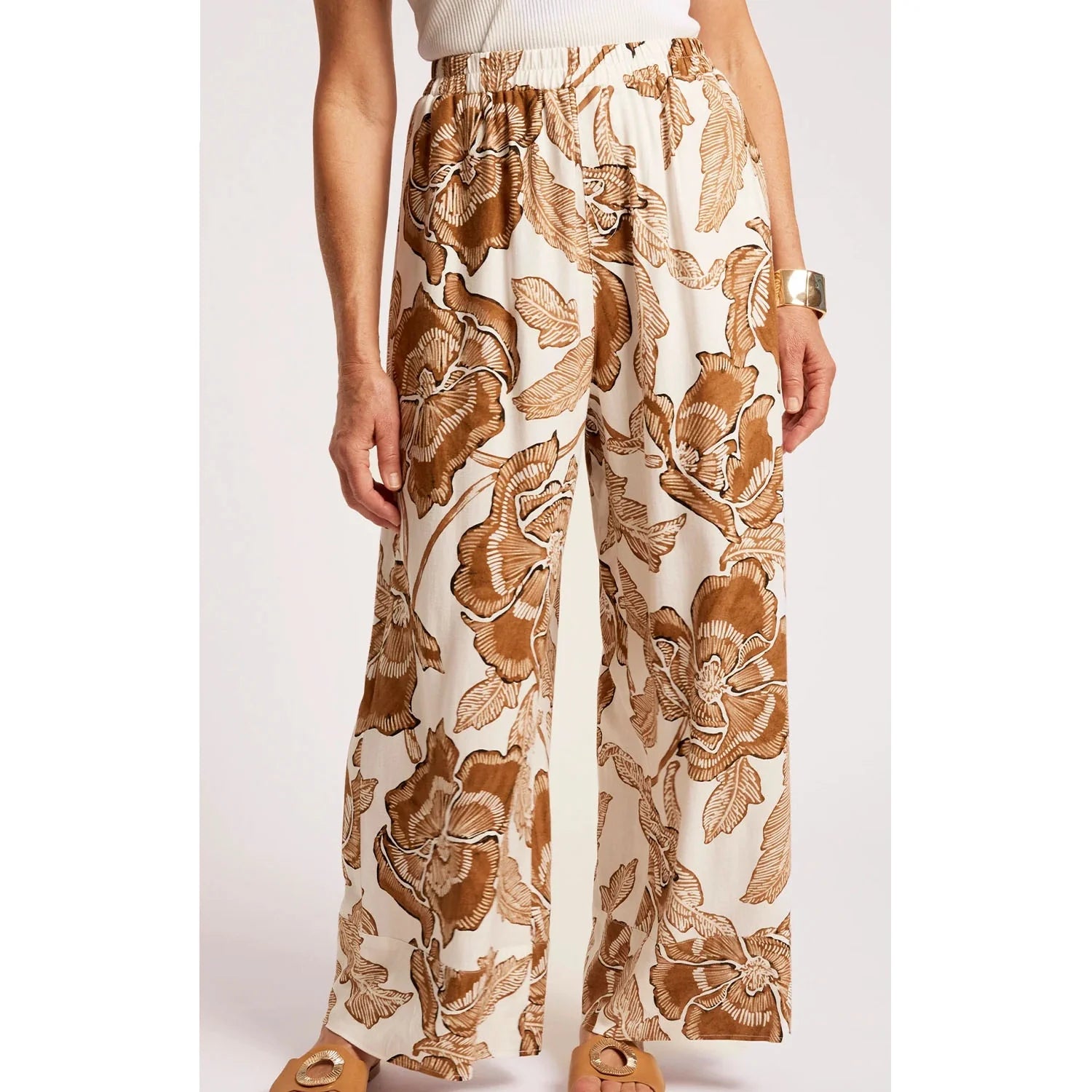 Image of A woman wearing Eb & Ive high-waisted, wide-leg pants with a brown and tan floral print on an off-white background, paired with a white ribbed top and tan flat sandals.