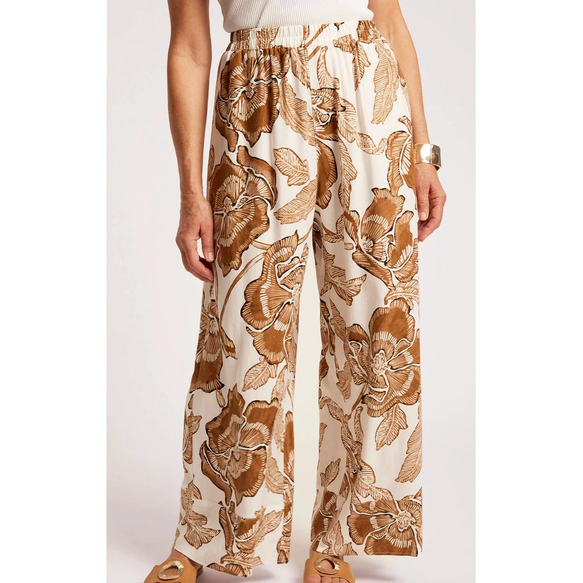 A woman wearing Eb & Ive high-waisted, wide-leg pants with a brown and tan floral print on an off-white background, paired with a white ribbed top and tan flat sandals.
