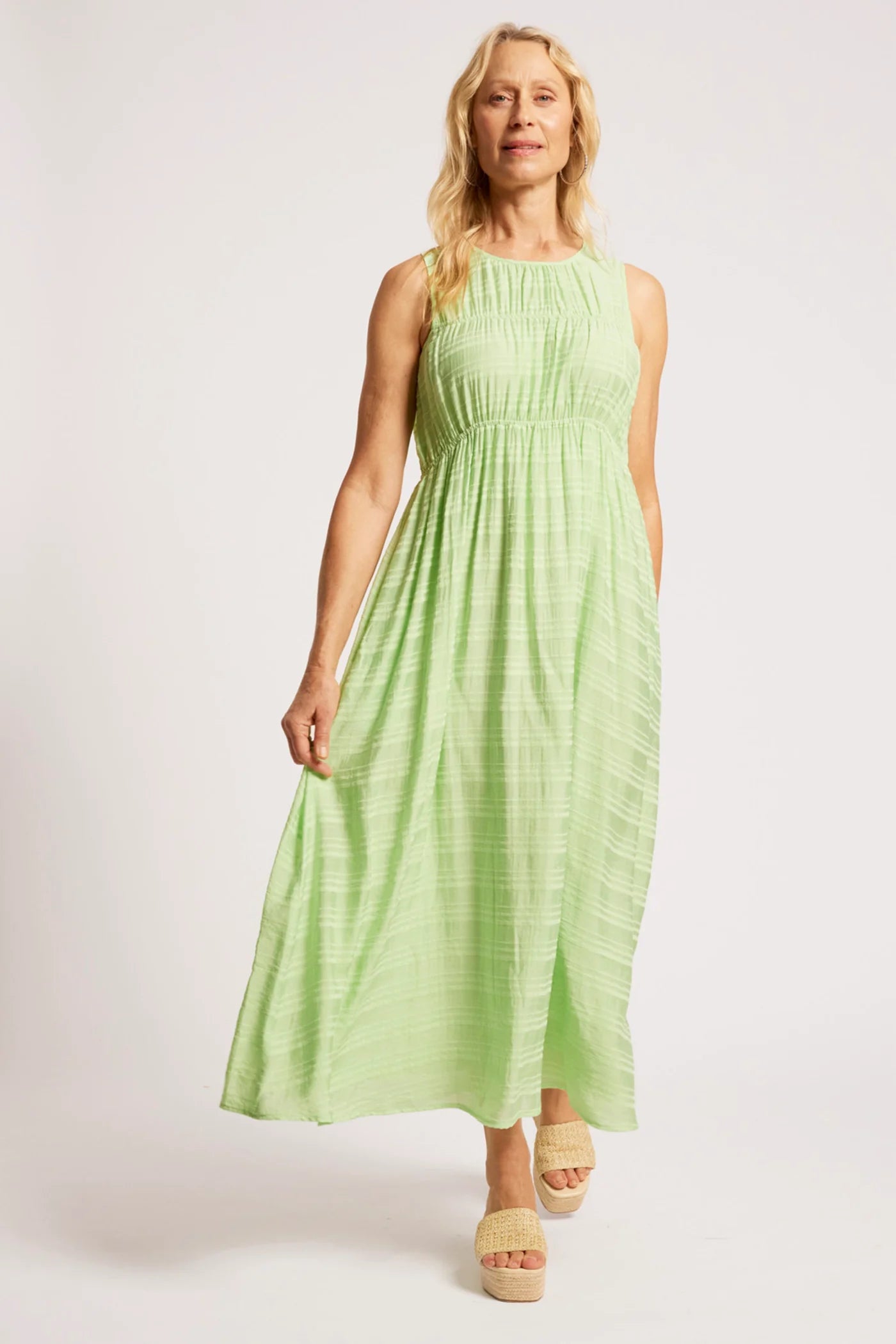 Image of A woman modeling a sleeveless Eb & Ive  light green maxi dress with a smocked bodice and gathered skirt, paired with beige platform sandals, against a plain light gray studio background.