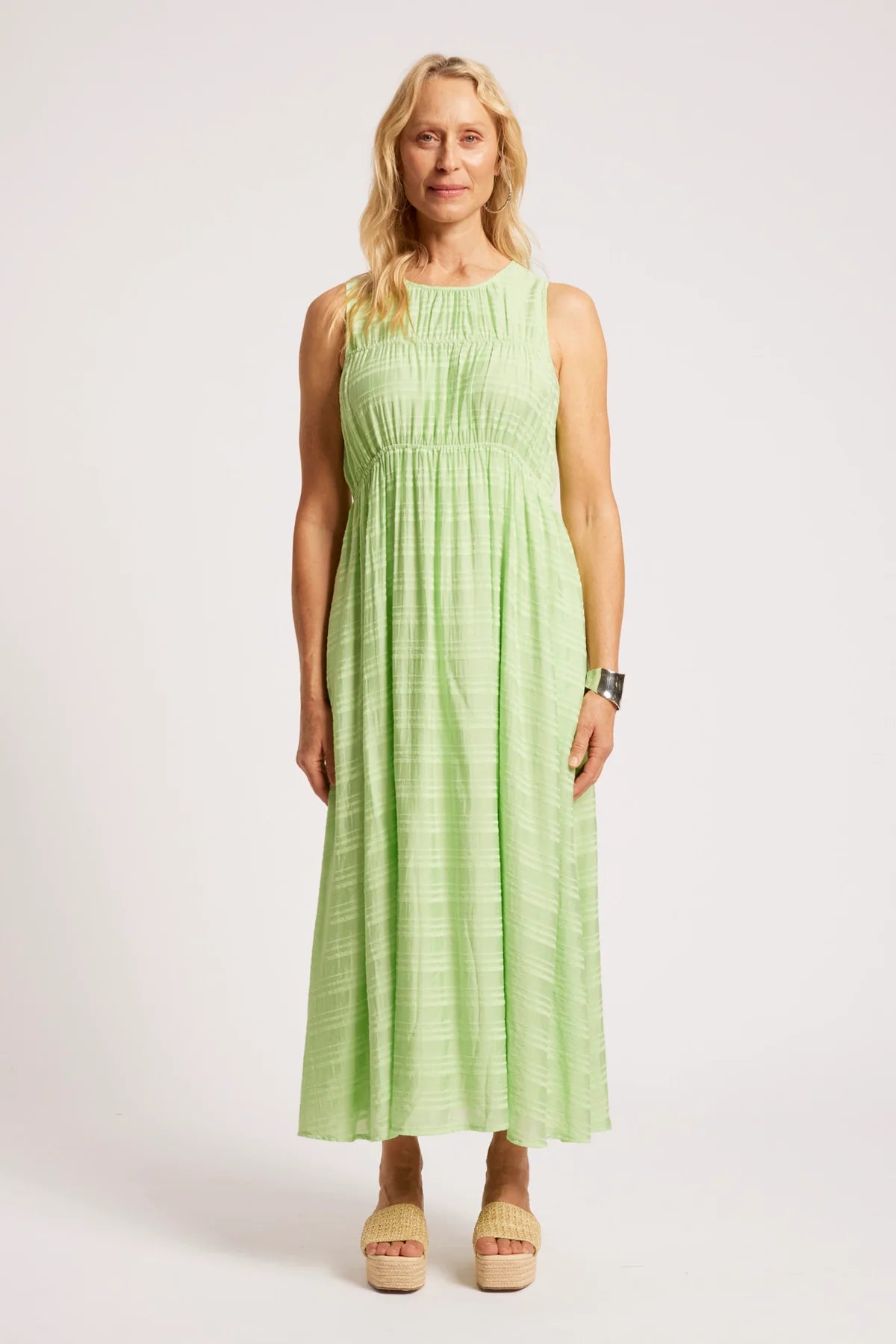 A woman modeling a Eb & Ive sleeveless light green maxi dress with a smocked bodice and textured fabric, paired with beige platform sandals, against a plain light gray studio background.