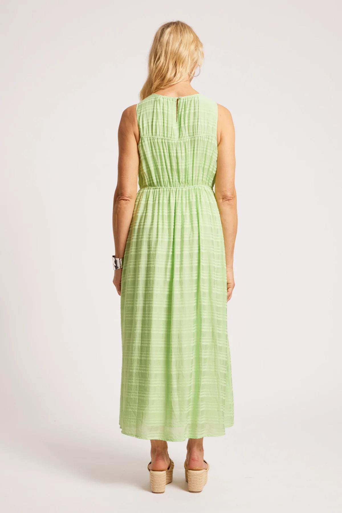 Image of A woman viewed from the back wearing a Eb & Ive sleeveless light green midi dress with a smocked back and pleated skirt, paired with beige wedge sandals, against a plain light gray studio background.