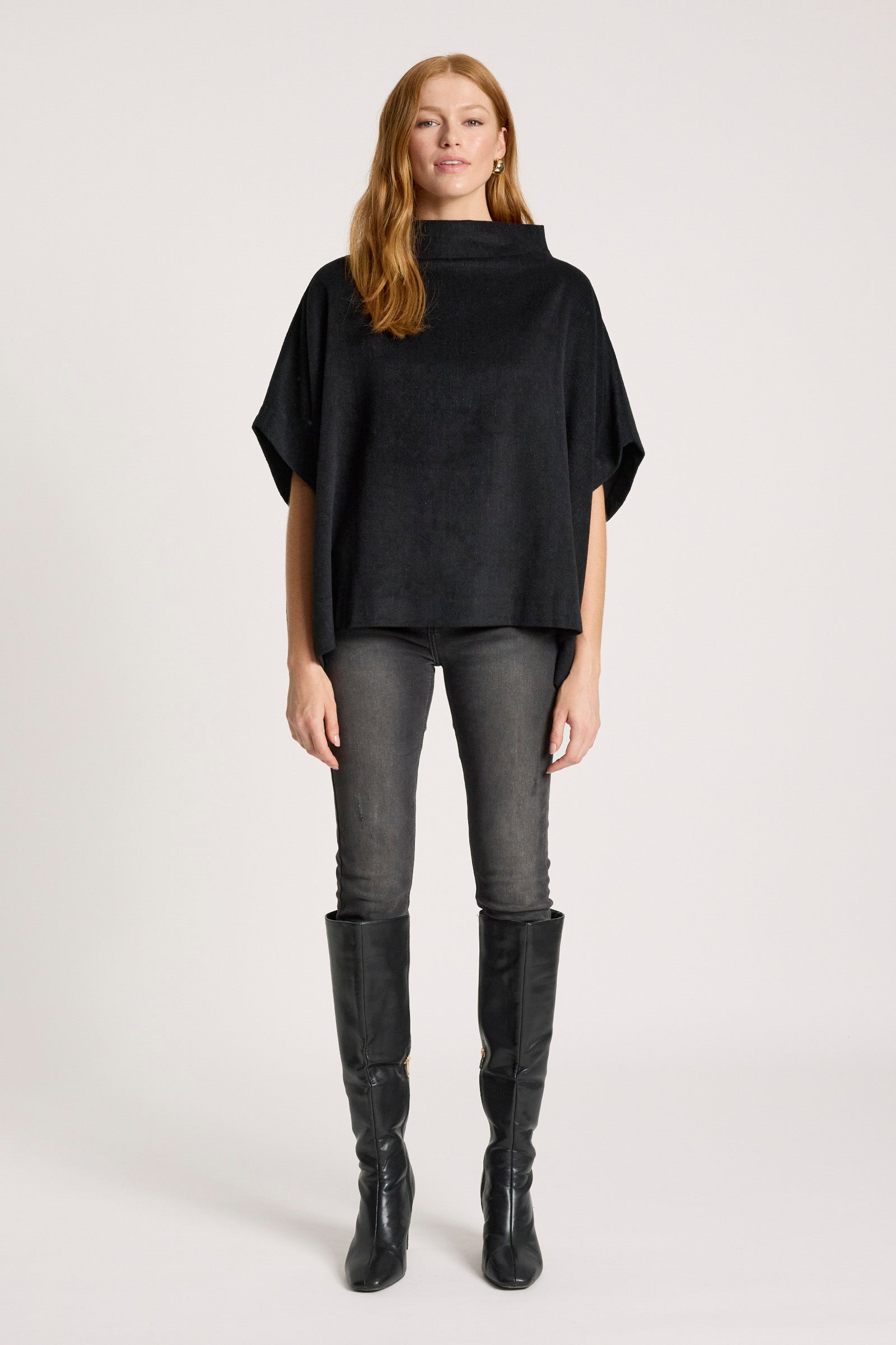 Woman wearing a Eb & Ive black poncho, grey leggings, and black knee-high boots on a white background