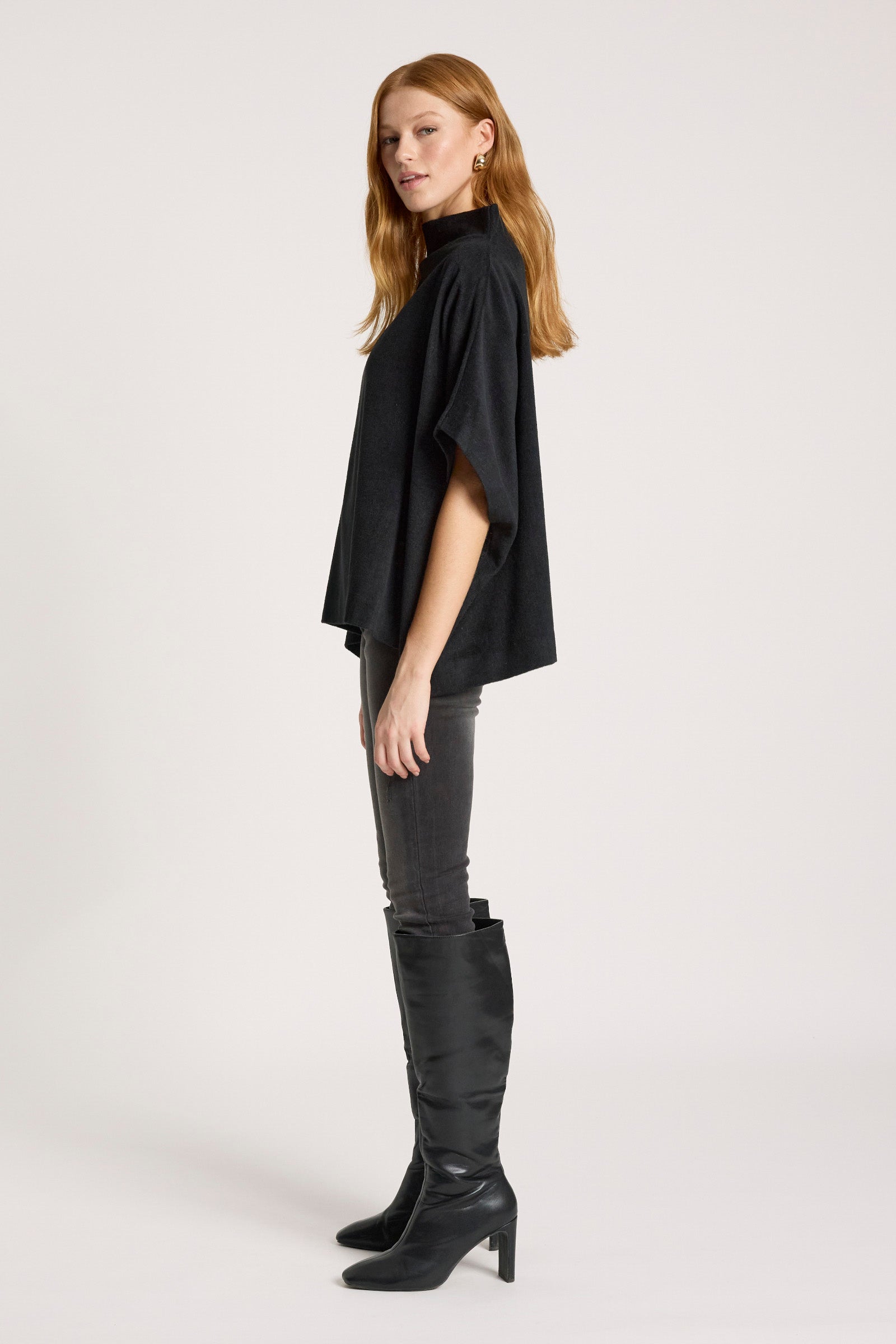 Woman wearing a Eb and Ive black poncho, gray pants, and black knee-high boots on a white background