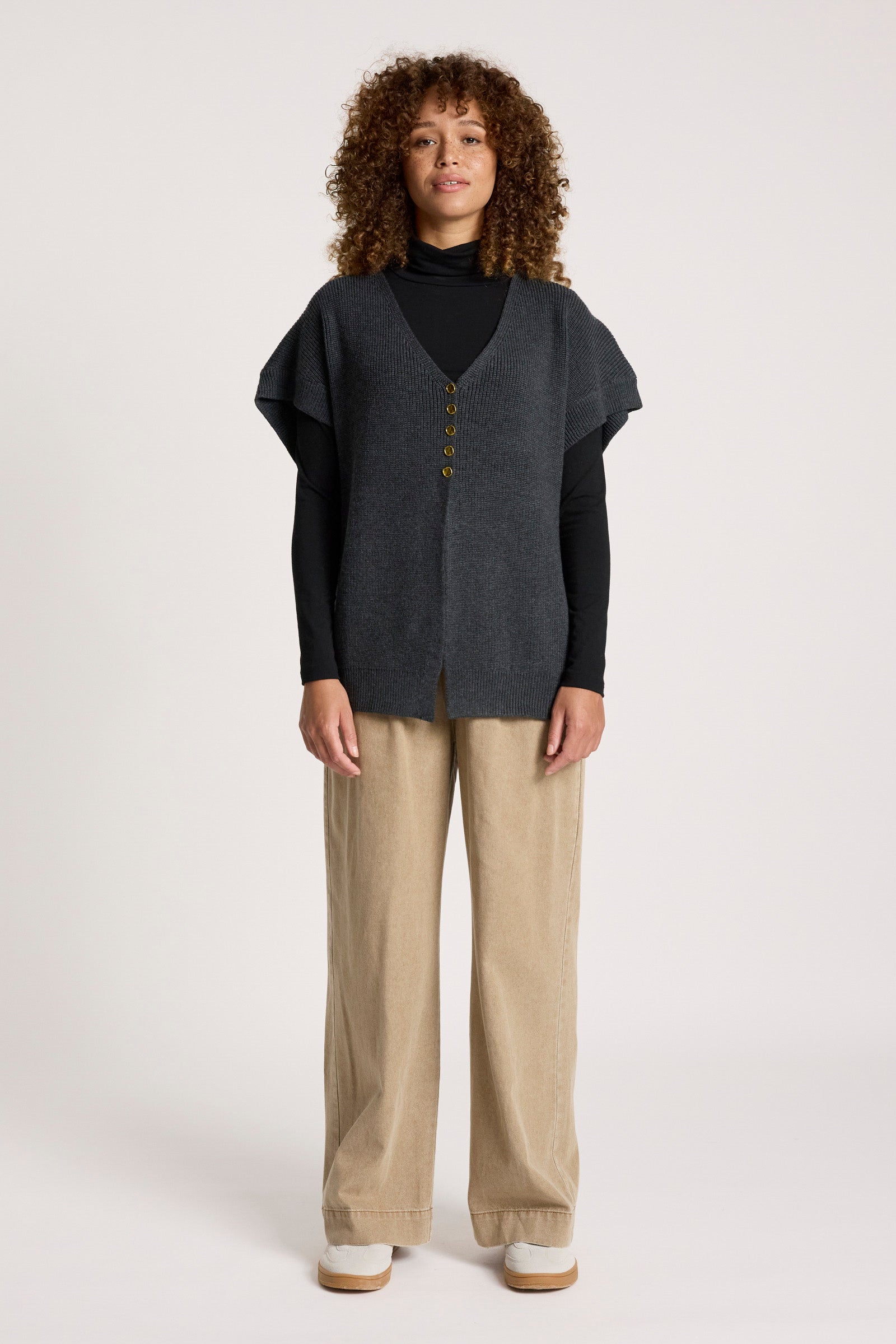 Image of Woman wearing a Eb & Ive dark grey vest over a black turtleneck with beige pants on a white background