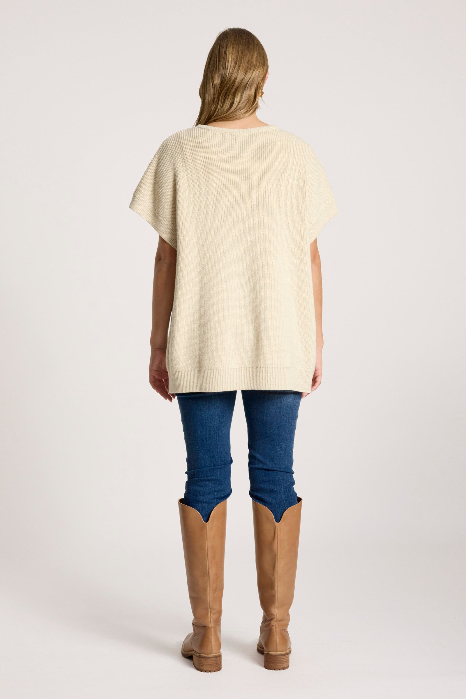 Woman wearing a Eb & Ive cream-colored top, blue pants, and brown boots on a white background