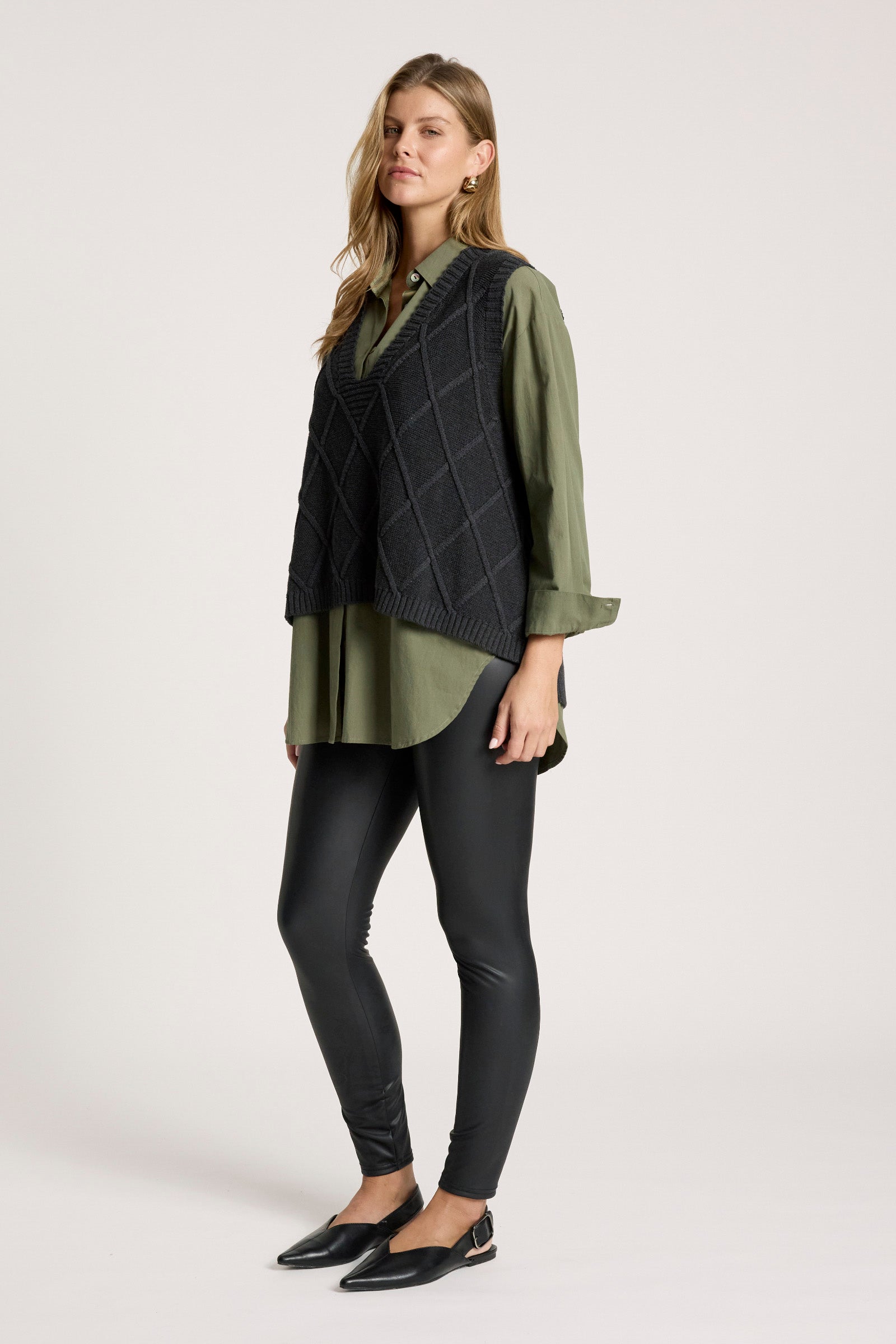 Image of Woman wearing a green shirt, Eb and Ive black vest, and black leggings on a white background