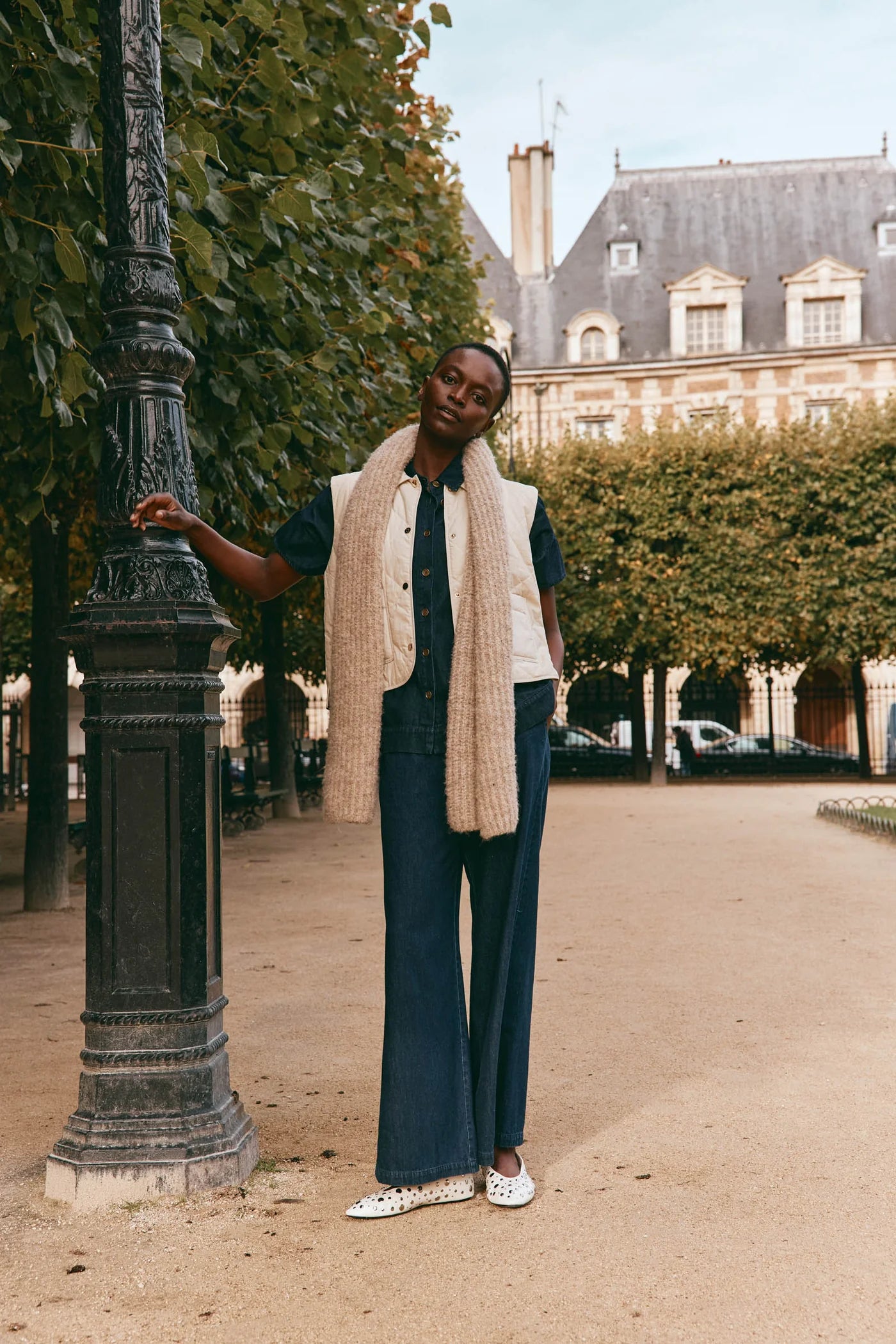 Image of Woman standing outdoors in a park-like setting with trees and a building in the background. Wearing eb & ive scarf