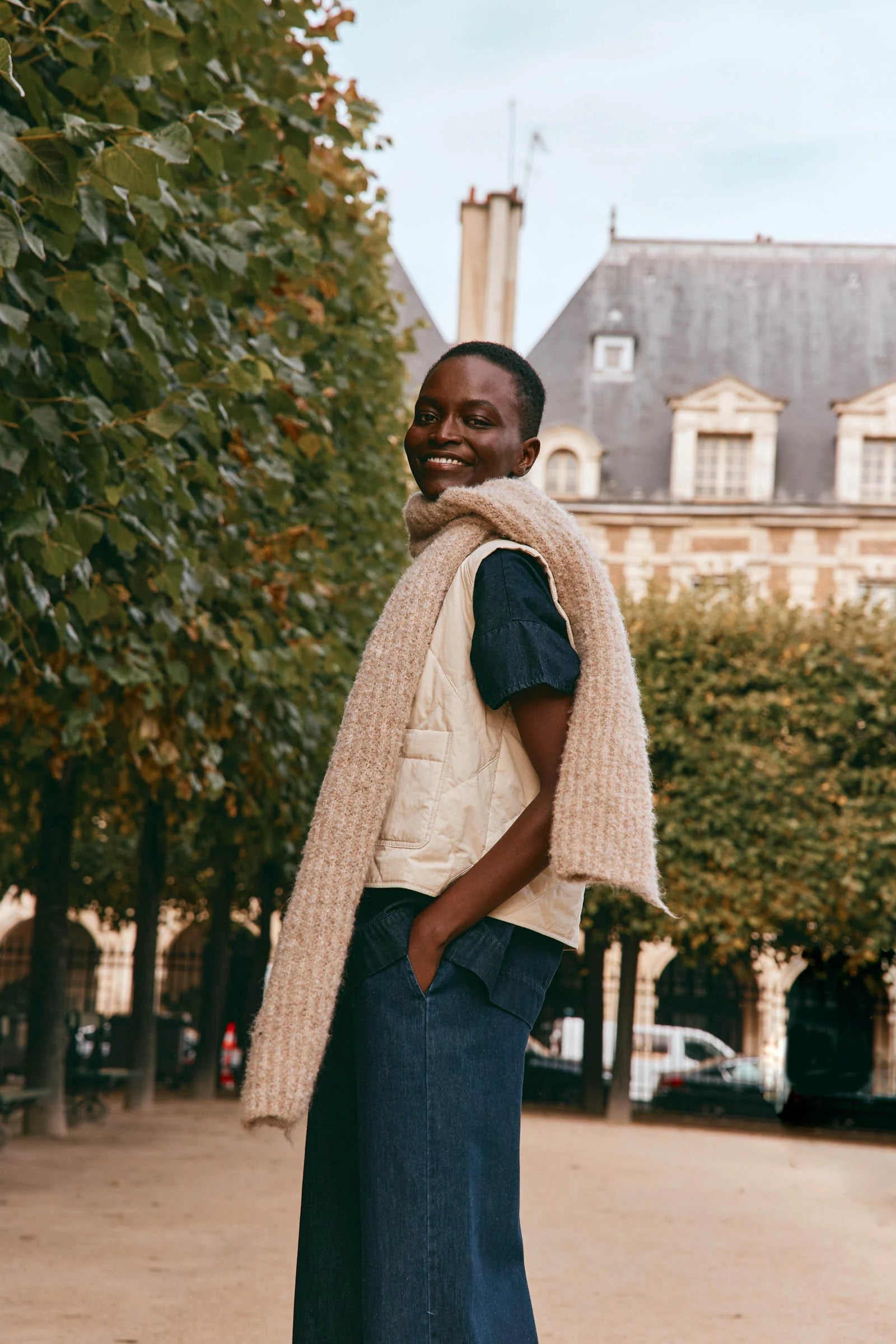Woman wearing a beige knitted sweater with a Eb & Ive scarf over a dark shirt in an outdoor setting with trees and buildings. 