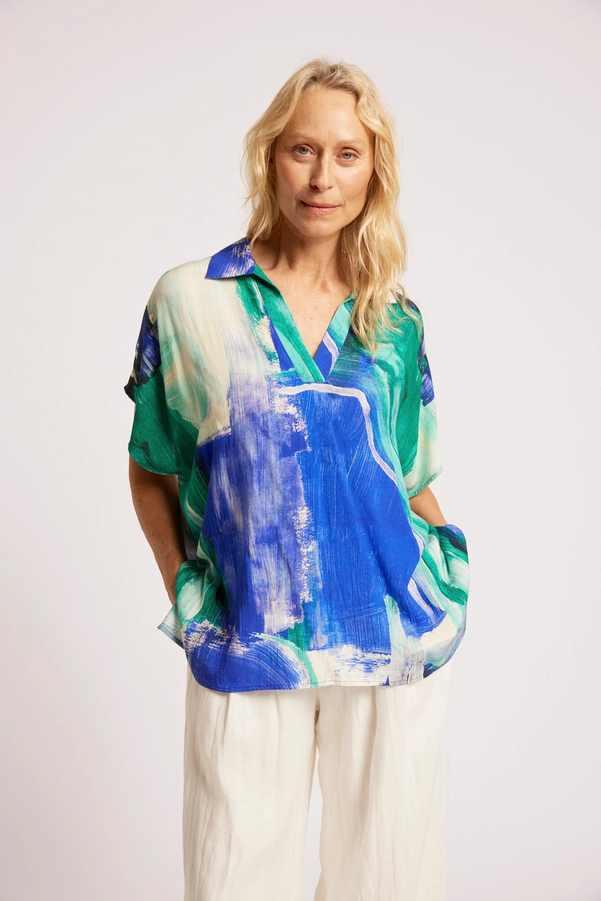 A woman modeling a Eb & Ive short-sleeve collared blouse with an abstract painterly print in blue, green, and white, paired with white trousers, against a plain light gray studio background.
