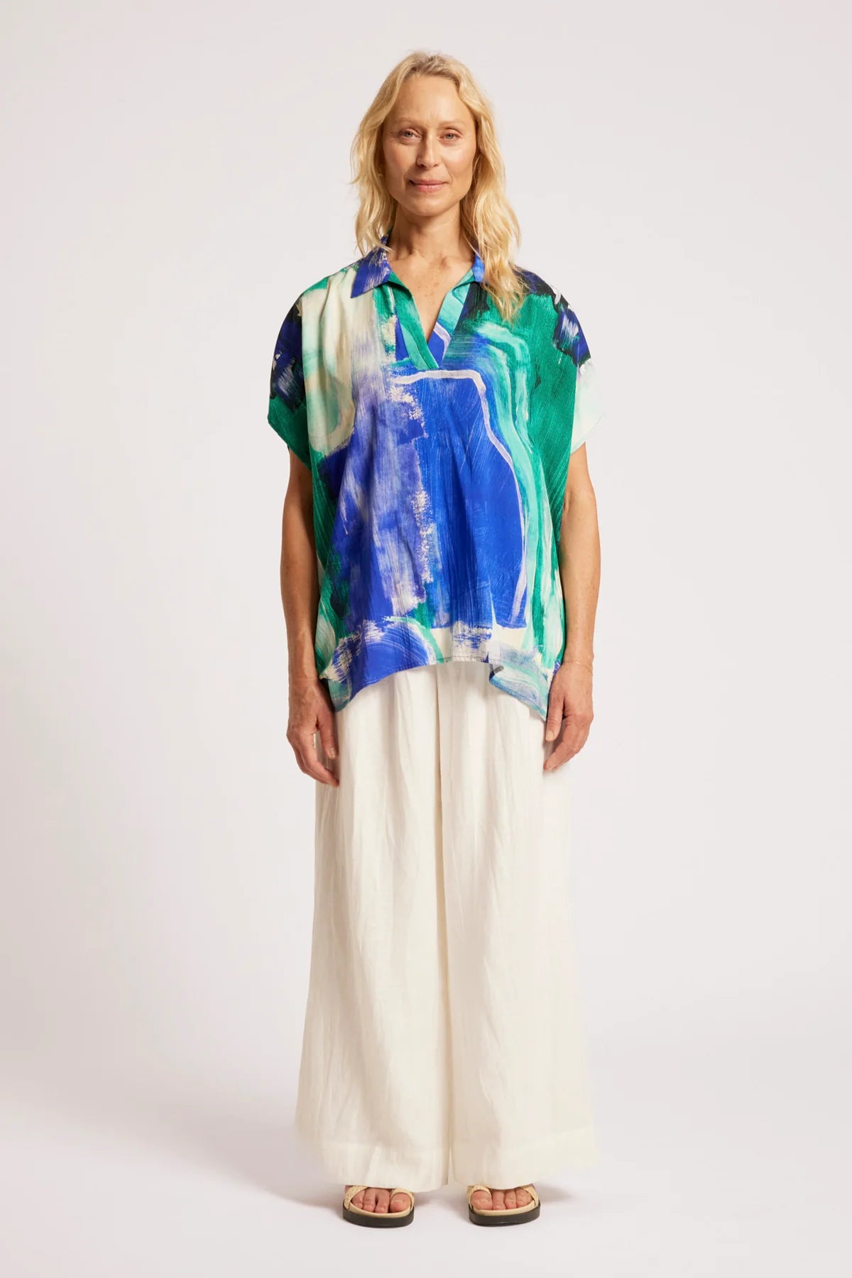 A woman stands against a plain light background wearing a Eb & Ive short-sleeve collared blouse with an abstract blue, green, and white print, paired with wide-leg cream pants and sandals.