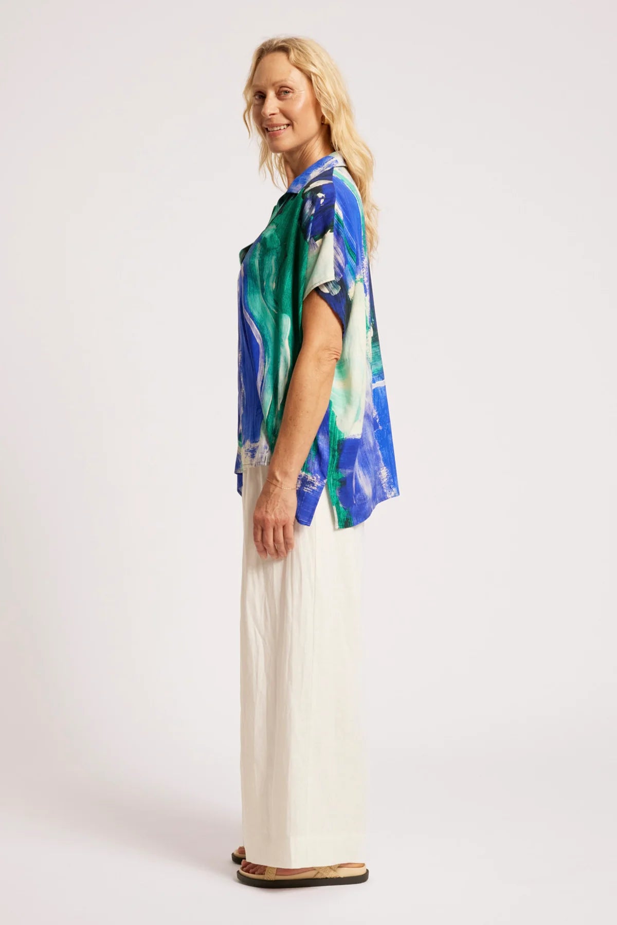 A woman modeling a Eb & Ive short-sleeve button-up shirt with a blue, green, and white abstract print, paired with white wide-leg pants and brown sandals, photographed against a plain light gray studio background.