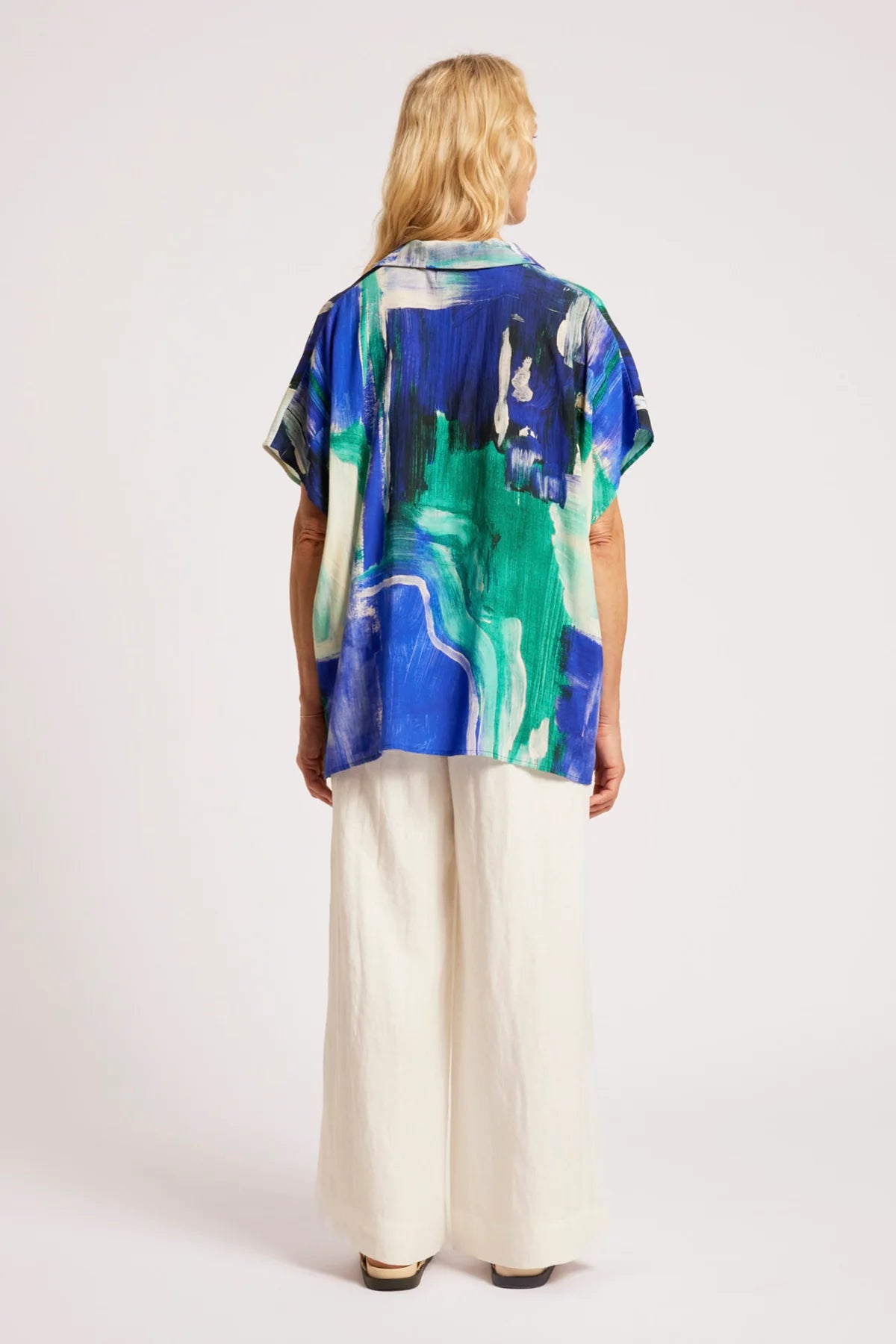 Image of A woman viewed from behind wearing a Eb & Ive short-sleeve button-up shirt with an abstract blue, green, and white print, paired with wide-leg cream trousers, against a plain light background.