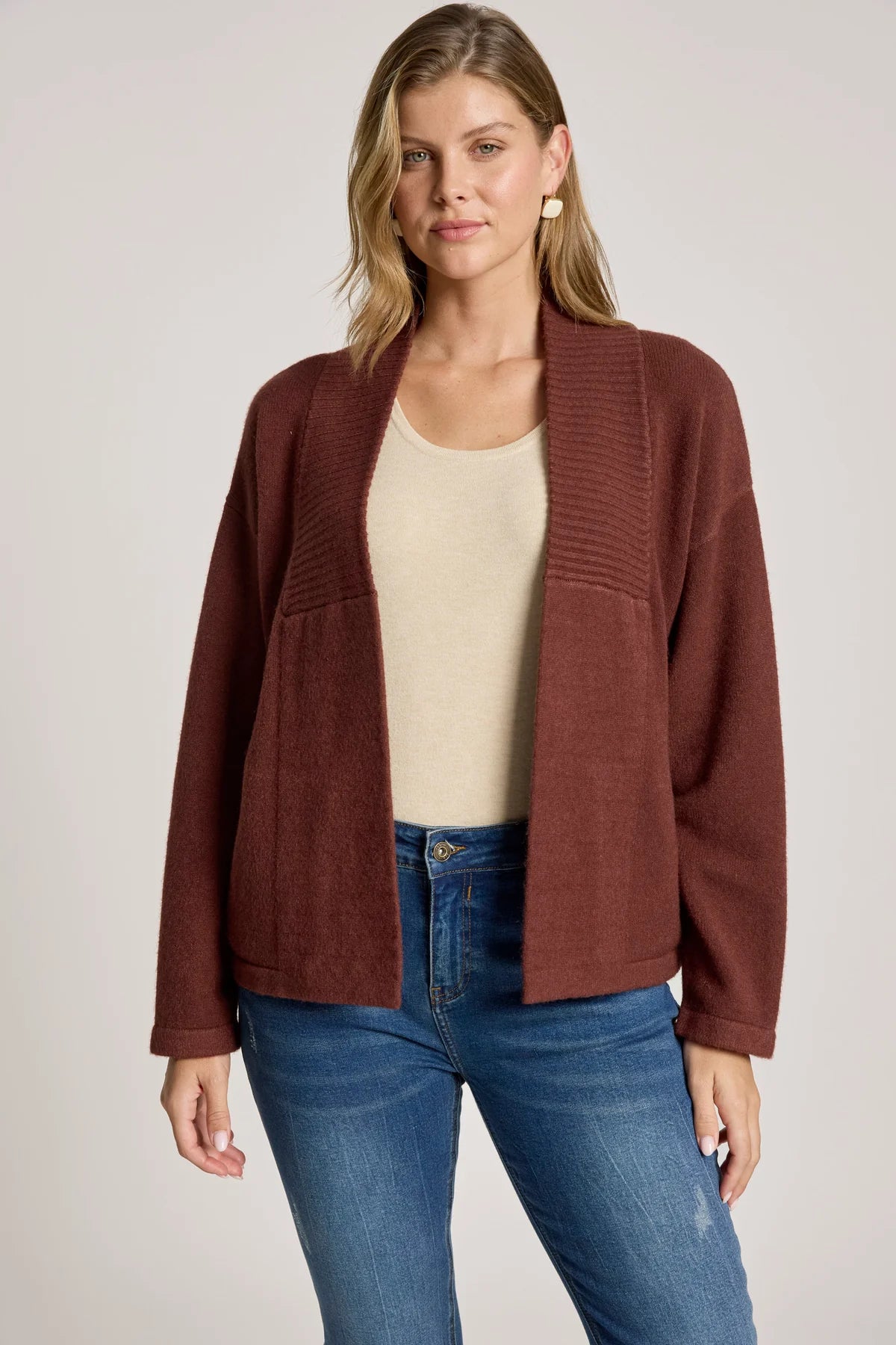Woman wearing a Eb & Ive brown cardigan over a beige top and blue jeans on a plain background