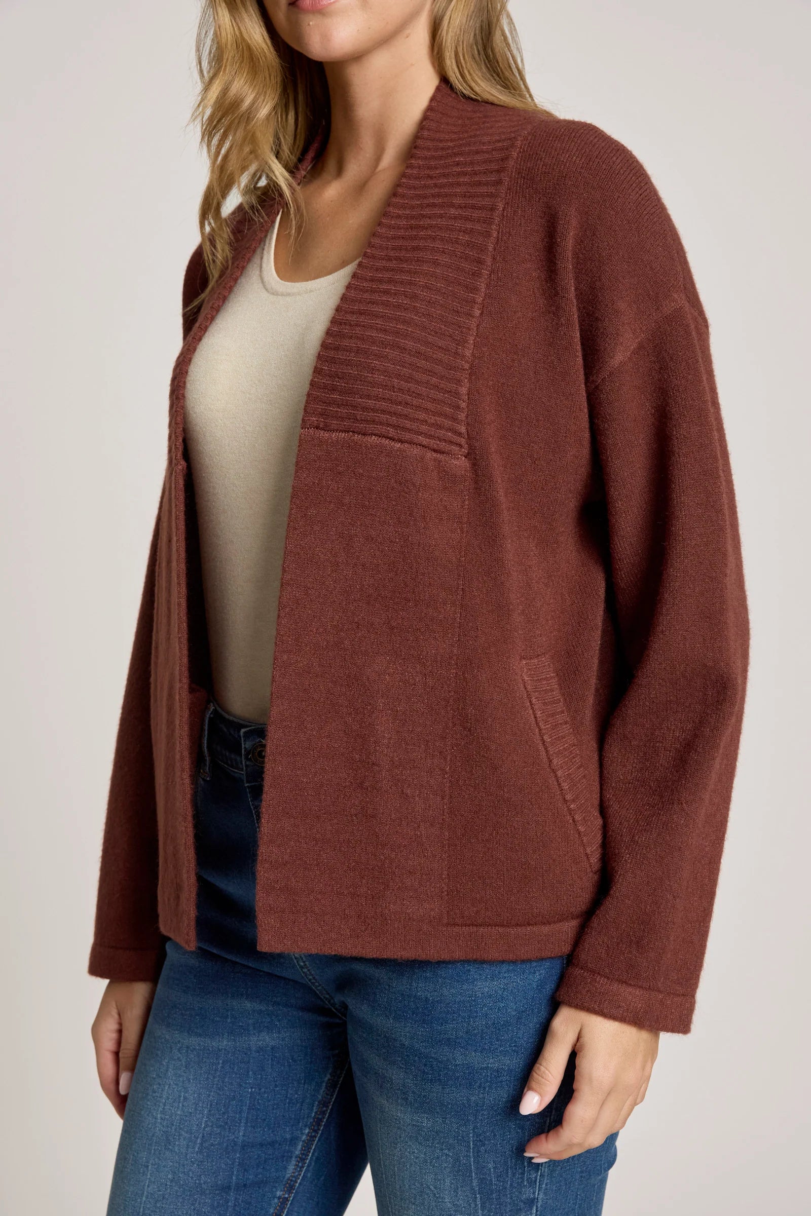 Woman wearing a Eb & Ive brown cardigan over a beige top and blue jeans on a plain background