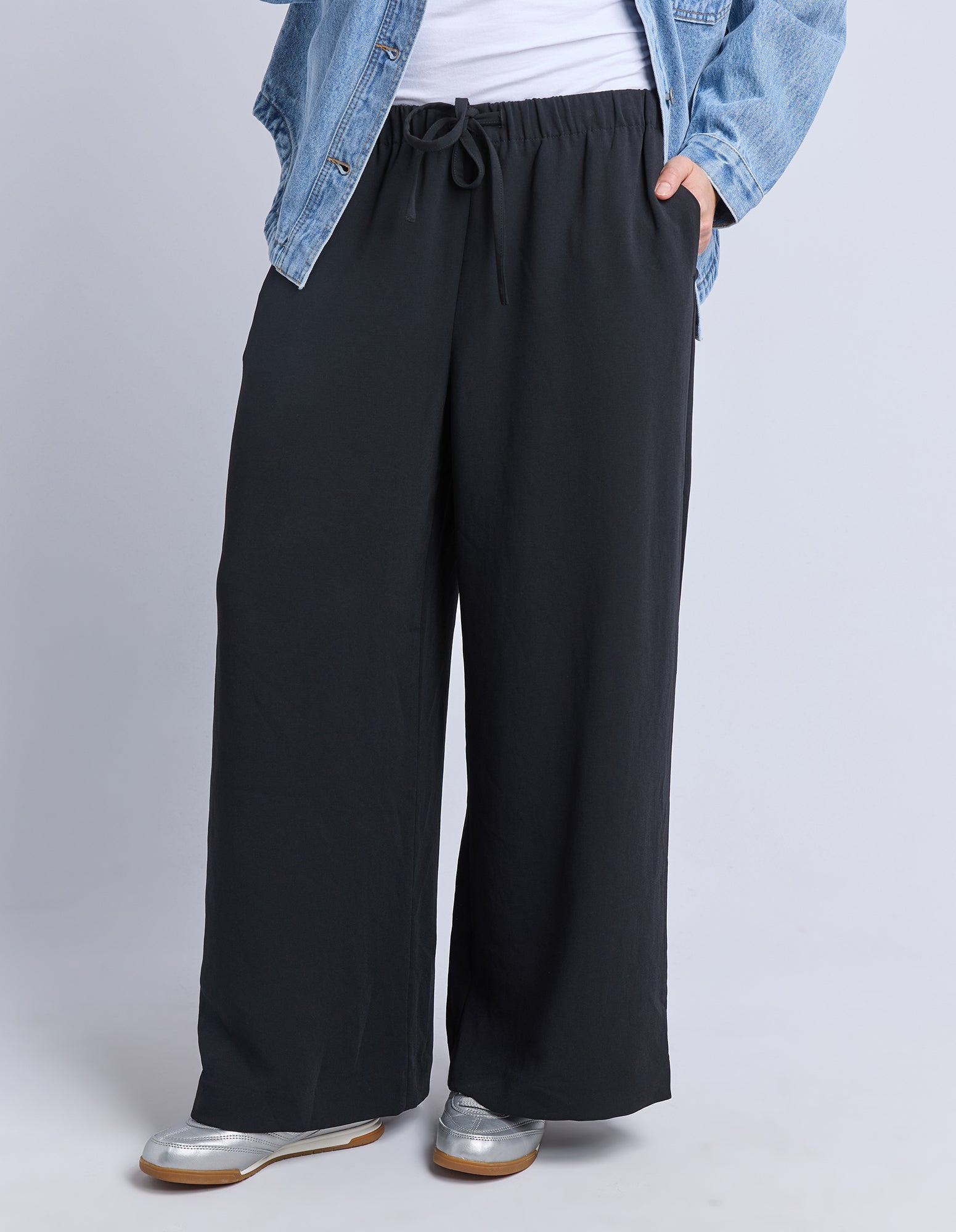 Woman wearing Elm black wide-leg pants with a denim jacket and white shirt on a light grae background