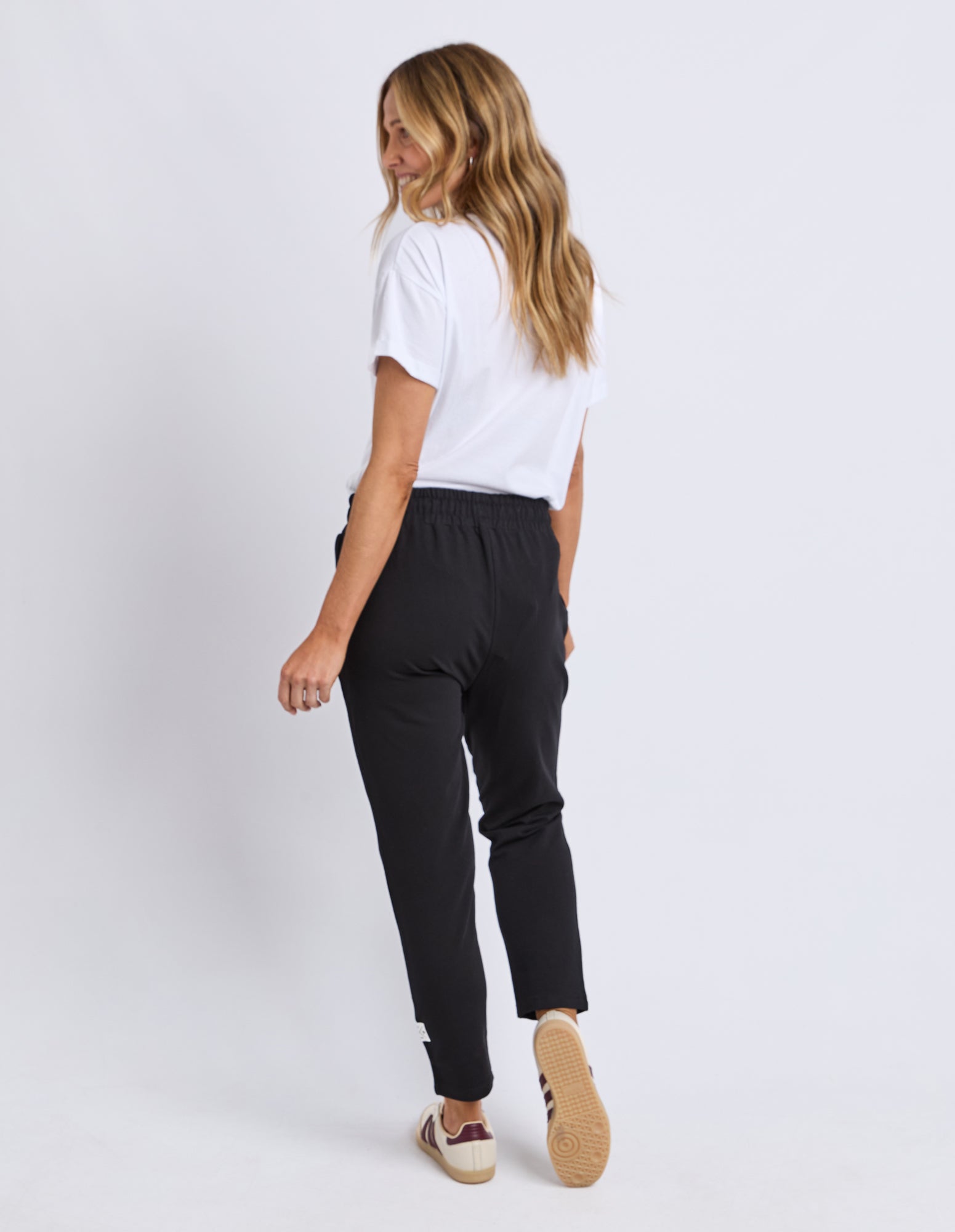 Back view of a Woman wearing a white t-shirt and Elm black Track pants on a plain background