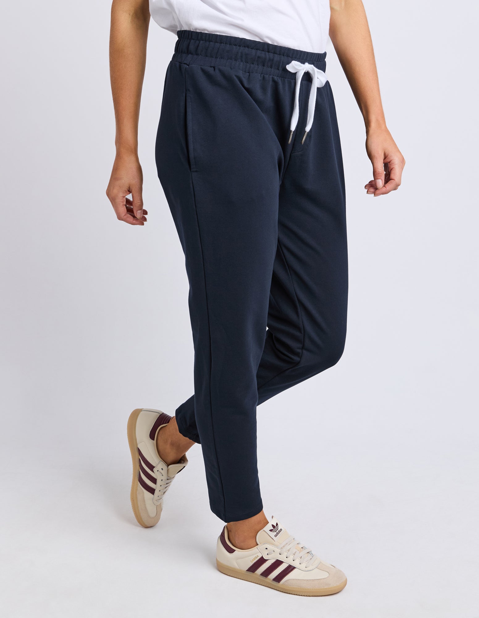 Side view of a woman wearing Elm navy track pants with a white drawstring, white shirt, and beige sneakers on a light grey background