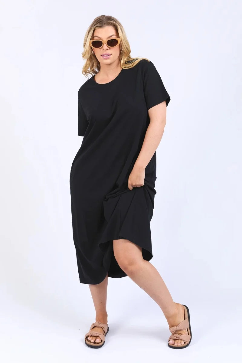 A woman modeling a Elm loose-fitting black short-sleeve midi dress with an asymmetrical draped hem, styled with tortoiseshell sunglasses and brown strappy sandals against a plain white studio background.