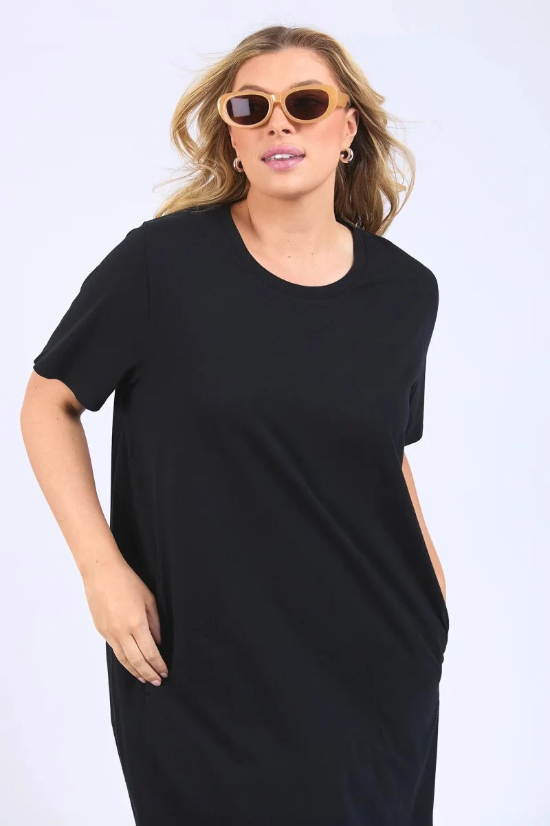 Image of A woman modeling a Elm loose-fitting black short-sleeve midi dress with an asymmetrical draped hem, styled with tortoiseshell sunglasses and brown strappy sandals against a plain white studio background.