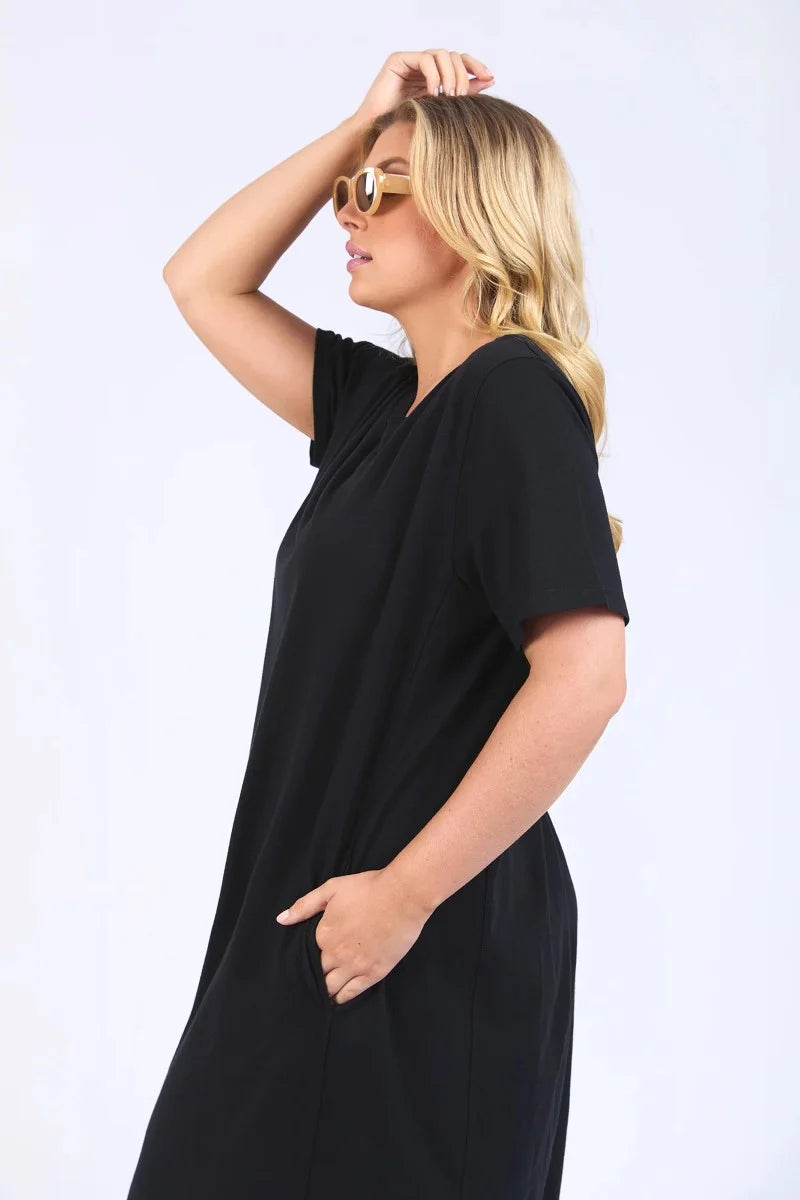 Image of A woman modeling a Elm loose-fitting black short-sleeve midi dress with an asymmetrical draped hem, styled with tortoiseshell sunglasses and brown strappy sandals against a plain white studio background.