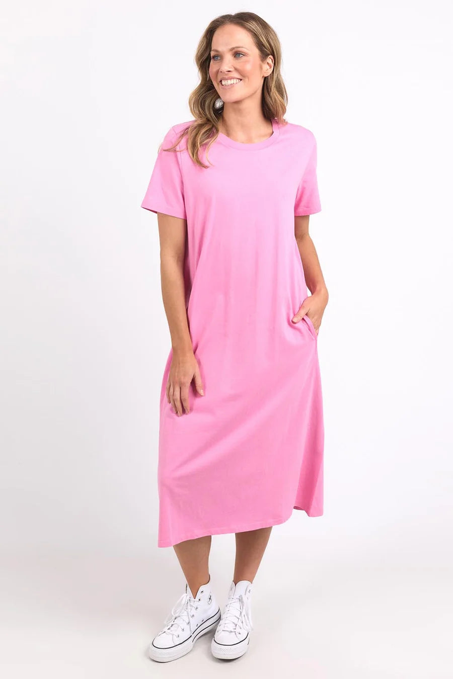 Image of A woman modeling a Elm pink short-sleeve midi dress with side pockets, paired with white sneakers, against a plain white studio background.