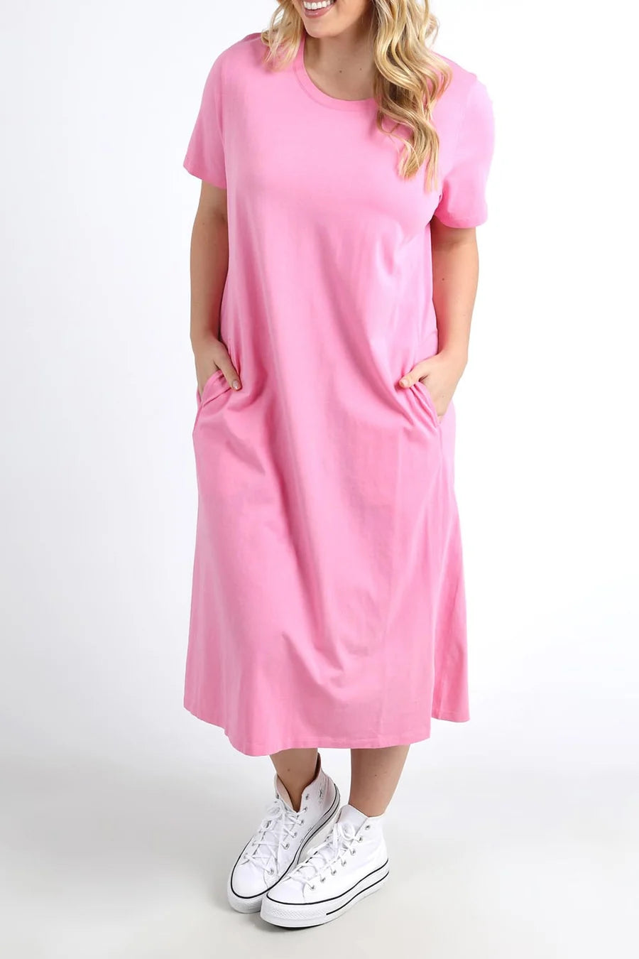 Image of A woman wearing a Elm pink short-sleeve midi dress with side pockets, paired with white sneakers, standing against a plain white studio background.