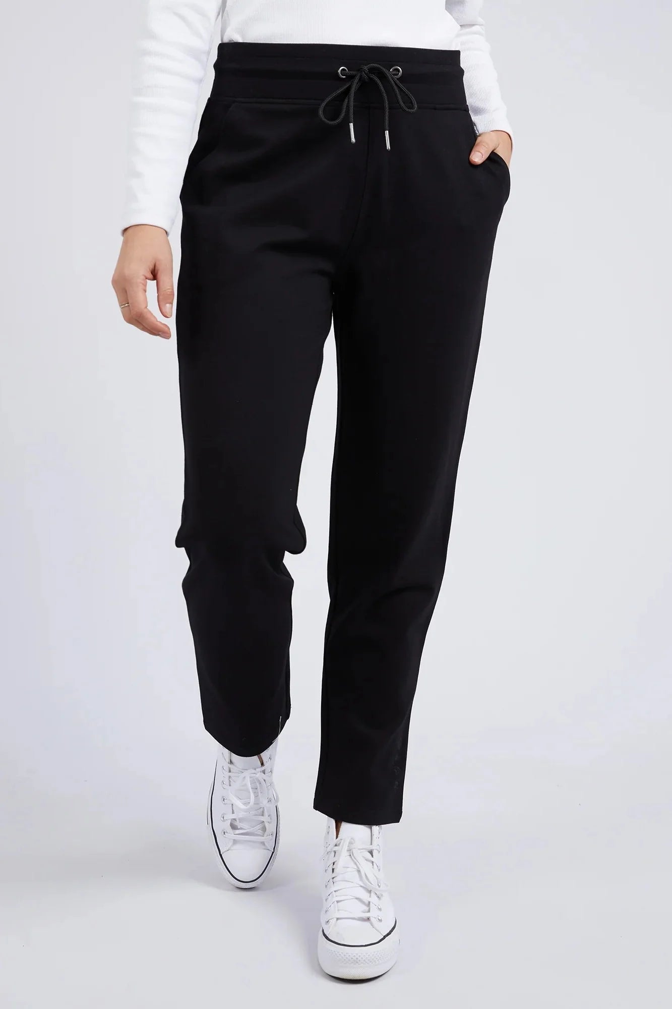 A woman wearing Elm black drawstring sweatpants with a white long-sleeve top and white sneakers, shown from the waist down against a light gray studio background.