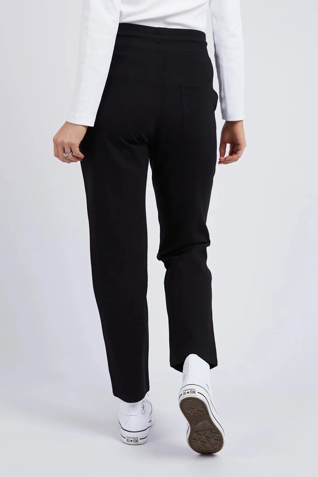 Image of A woman shown from the back wearing Elm black straight-leg trousers, a white long-sleeve top, and white sneakers against a light gray studio background.