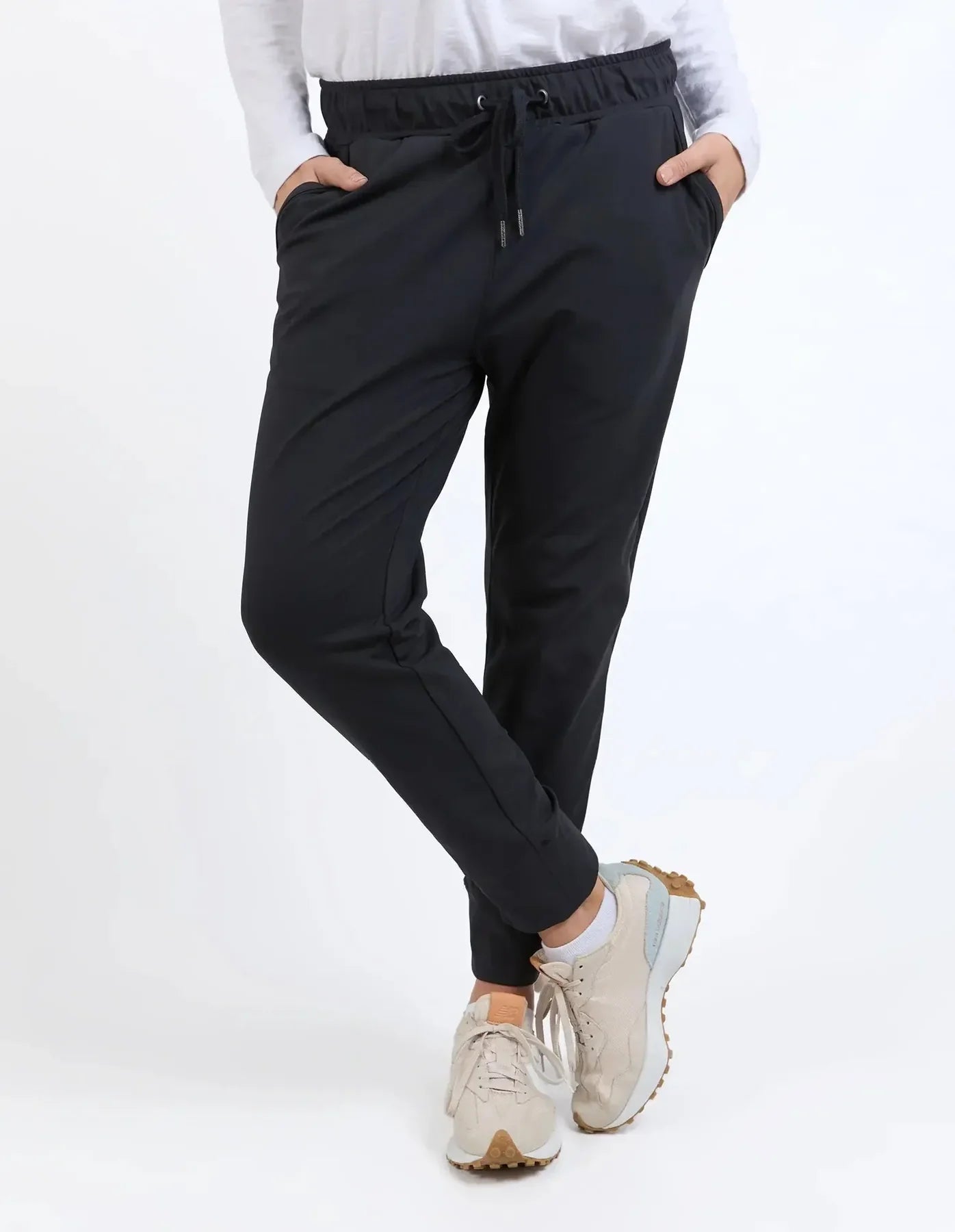 A woman wearing Elm black drawstring jogger-style pants with a white long-sleeve top and beige sneakers, standing against a plain light background.