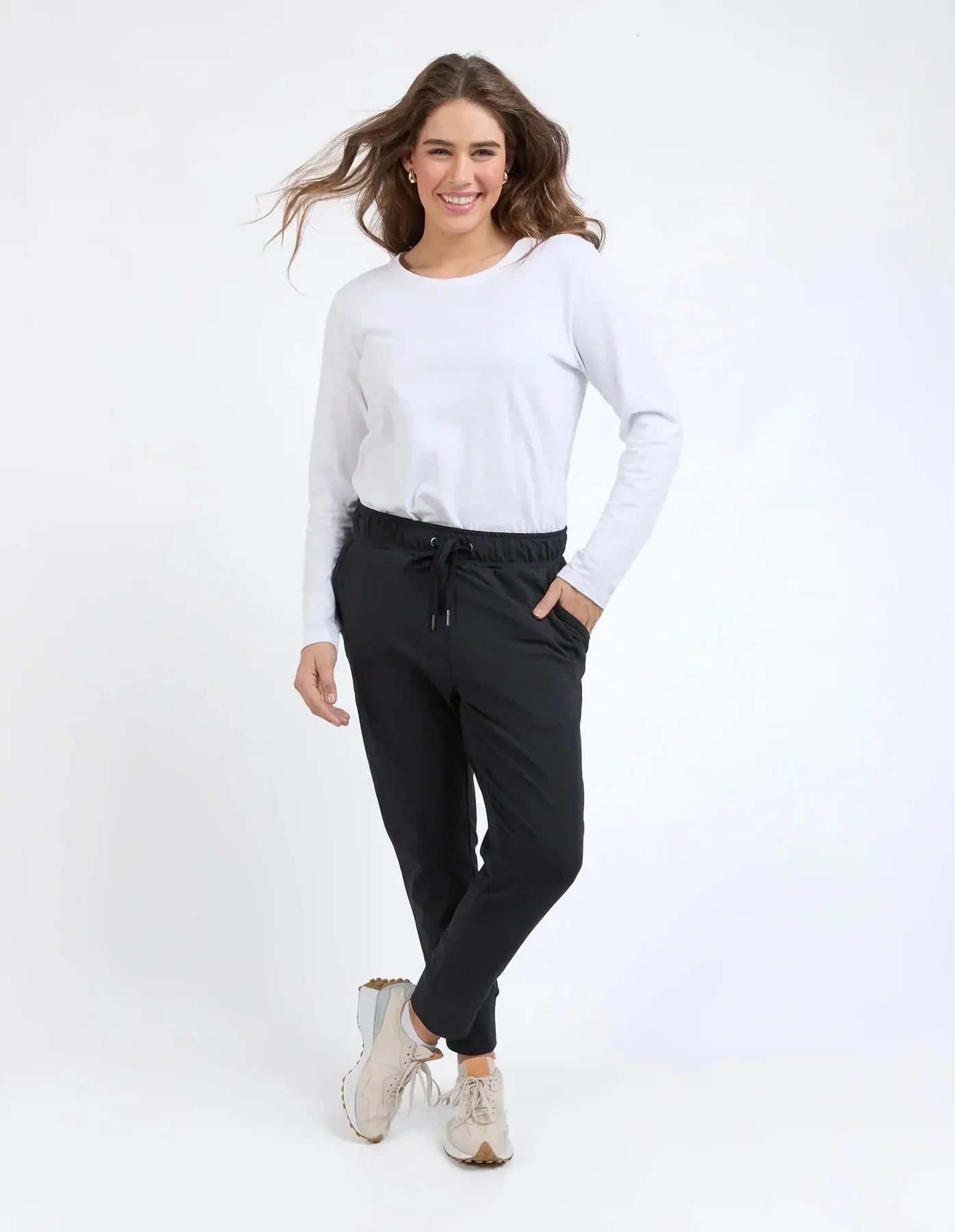 A woman modeling a white long-sleeve crewneck top and Elm black drawstring jogger pants with beige sneakers, standing against a plain white studio background.