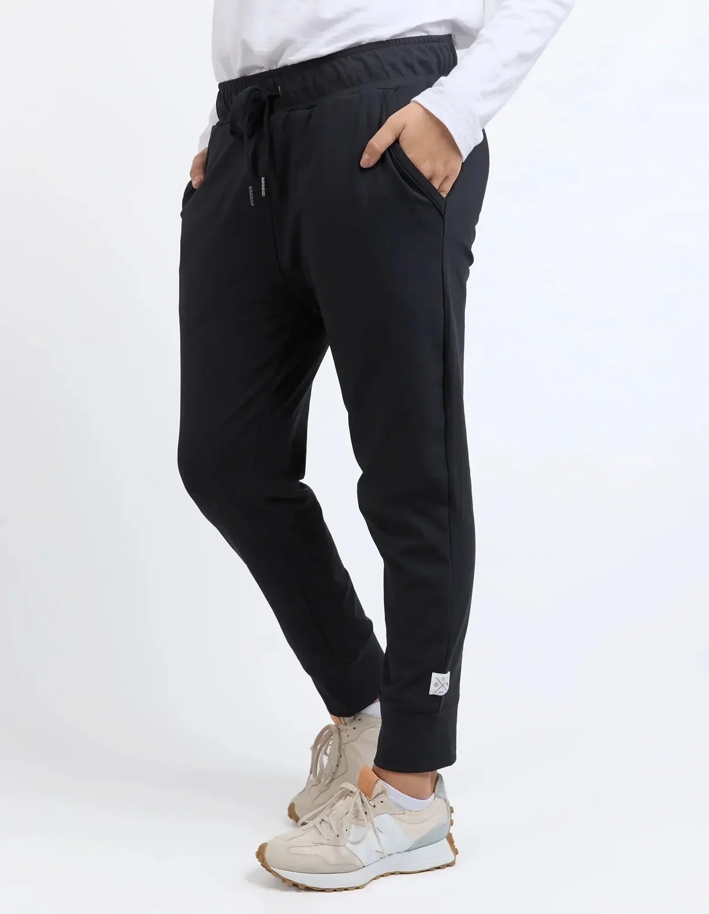 A woman wearing Elm black jogger-style sweatpants with a drawstring waist, side pockets, and a small white label near the cuff, paired with a white top and beige sneakers against a light gray studio background.