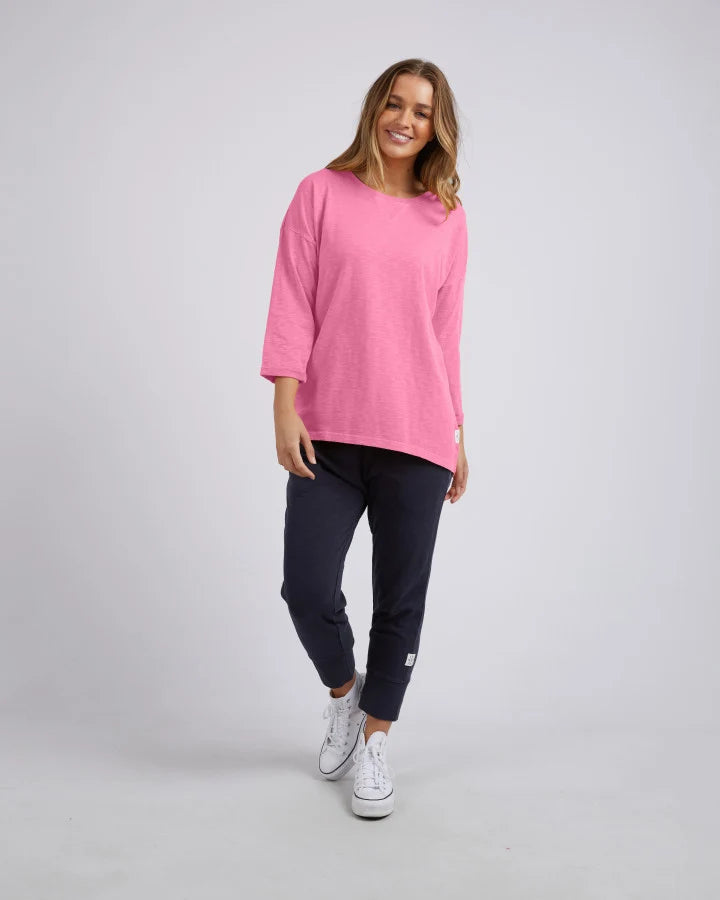 Image of A woman modeling a Elm bright pink oversized crewneck top, paired with dark navy pants and white sneakers, against a light gray studio background.