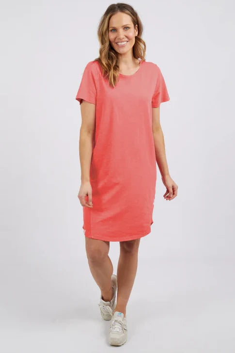 Image of A woman modeling a Elm coral pink short-sleeve t-shirt dress with side slits, paired with white sneakers, against a light gray studio background.