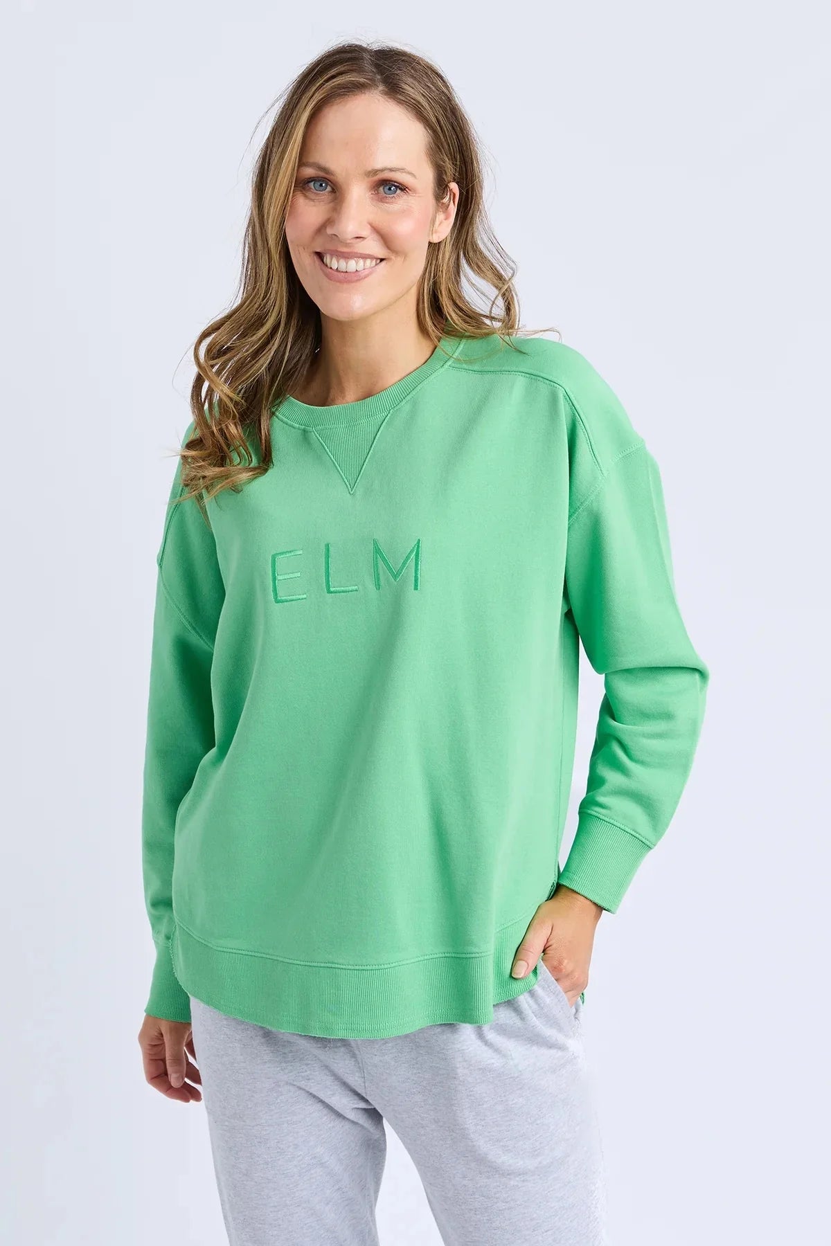 A woman wearing a Elm bright green crewneck sweatshirt with 'ELM' embroidered on the chest, paired with light gray sweatpants, against a light gray studio background.