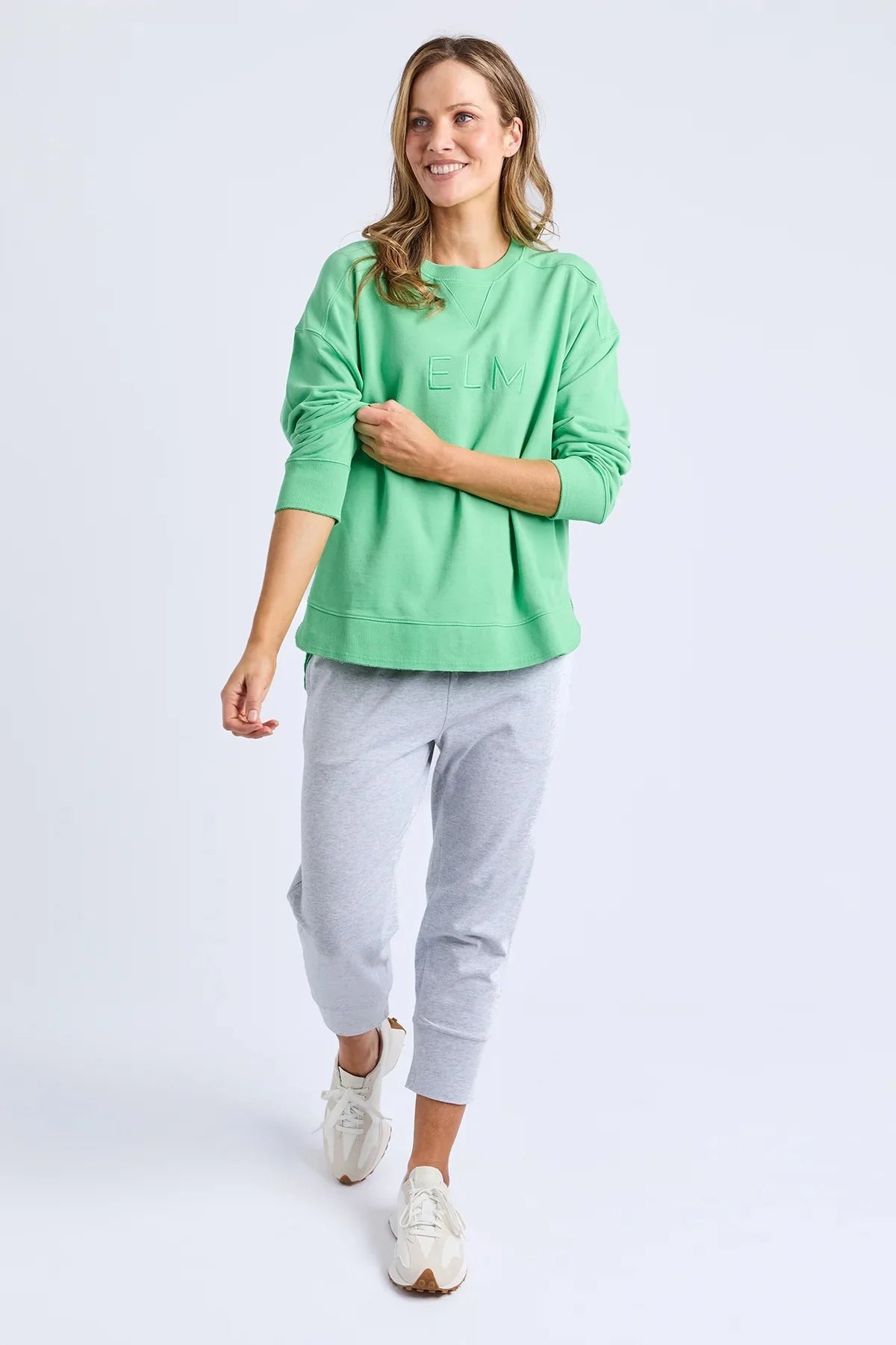 Image of A woman modeling a Elm mint green crewneck sweatshirt and light gray jogger pants with white sneakers, set against a plain light gray studio background.