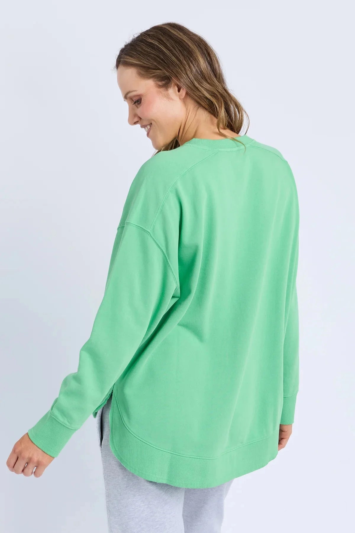 A woman wearing a Elm mint green long-sleeve crewneck sweatshirt, shown from a back-three-quarter angle against a light gray studio background.
