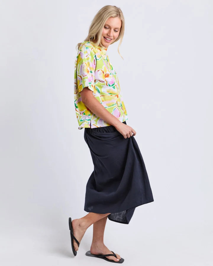A woman modeling a short-sleeve button-up shirt with a colorful tropical fruit print in yellow, green, pink, and orange, paired with a Elm dark navy midi skirt and black flip-flops, against a light gray studio background.