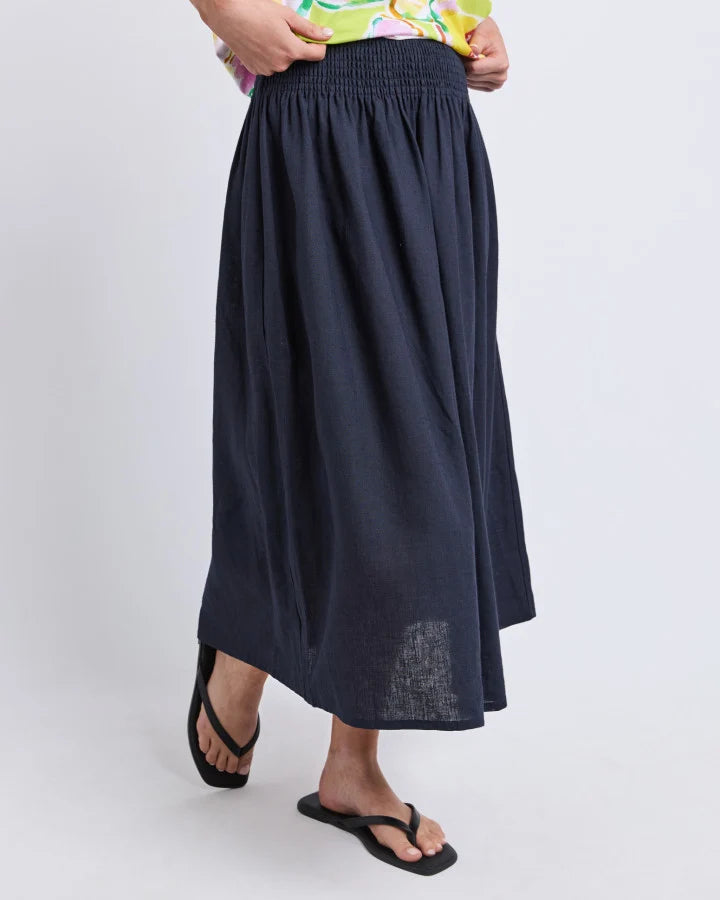 Image of A woman wearing a Elm dark navy blue linen midi skirt with a smocked elastic waistband, paired with black flip-flop sandals and a colorful top, shown against a light gray studio background.