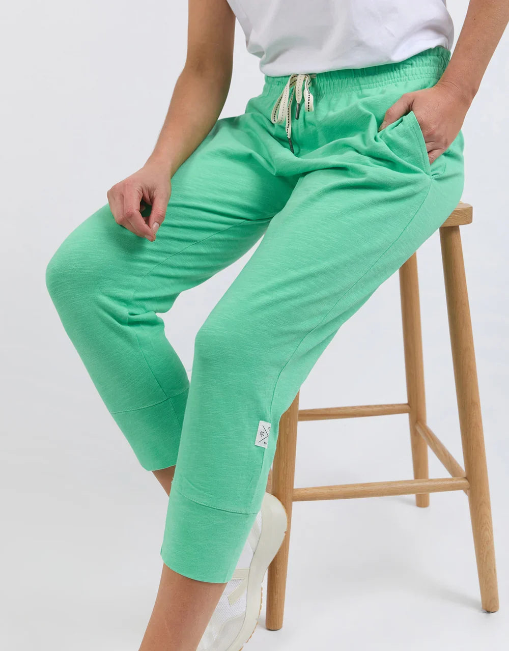 Image of A woman wearing Elm bright mint green cropped sweatpants with an elastic drawstring waist and side pockets, seated on a wooden stool against a light gray studio background.