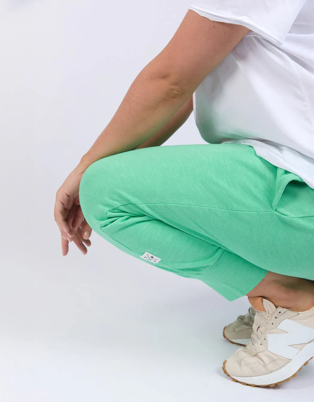 A woman crouching while wearing Elm mint green jogger pants, a white t-shirt, and beige sneakers against a plain white studio background.
