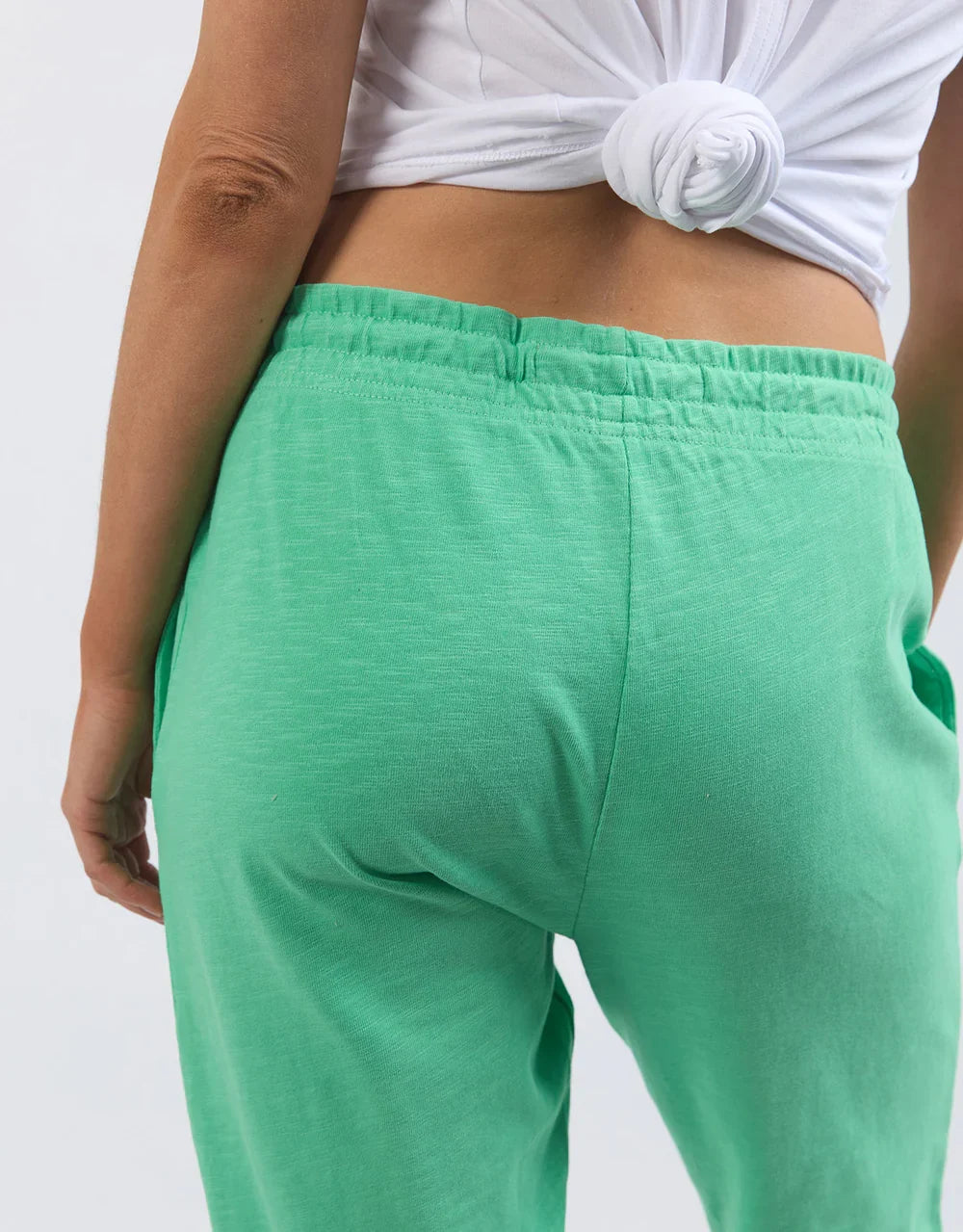 A close-up back view of a woman wearing Elm mint green lounge pants and a white knotted crop top against a plain light background.
