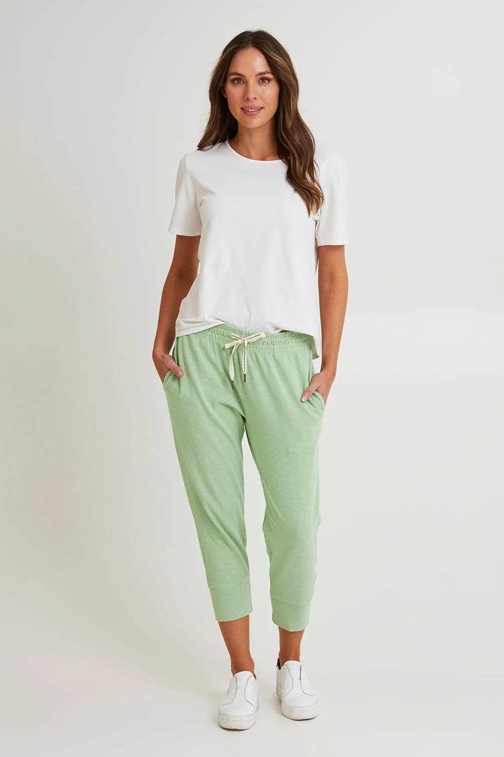 Image of A woman modeling a white short-sleeve t-shirt and Elm light sage green cropped jogger pants with a white drawstring, paired with white sneakers, against a plain light gray studio background.