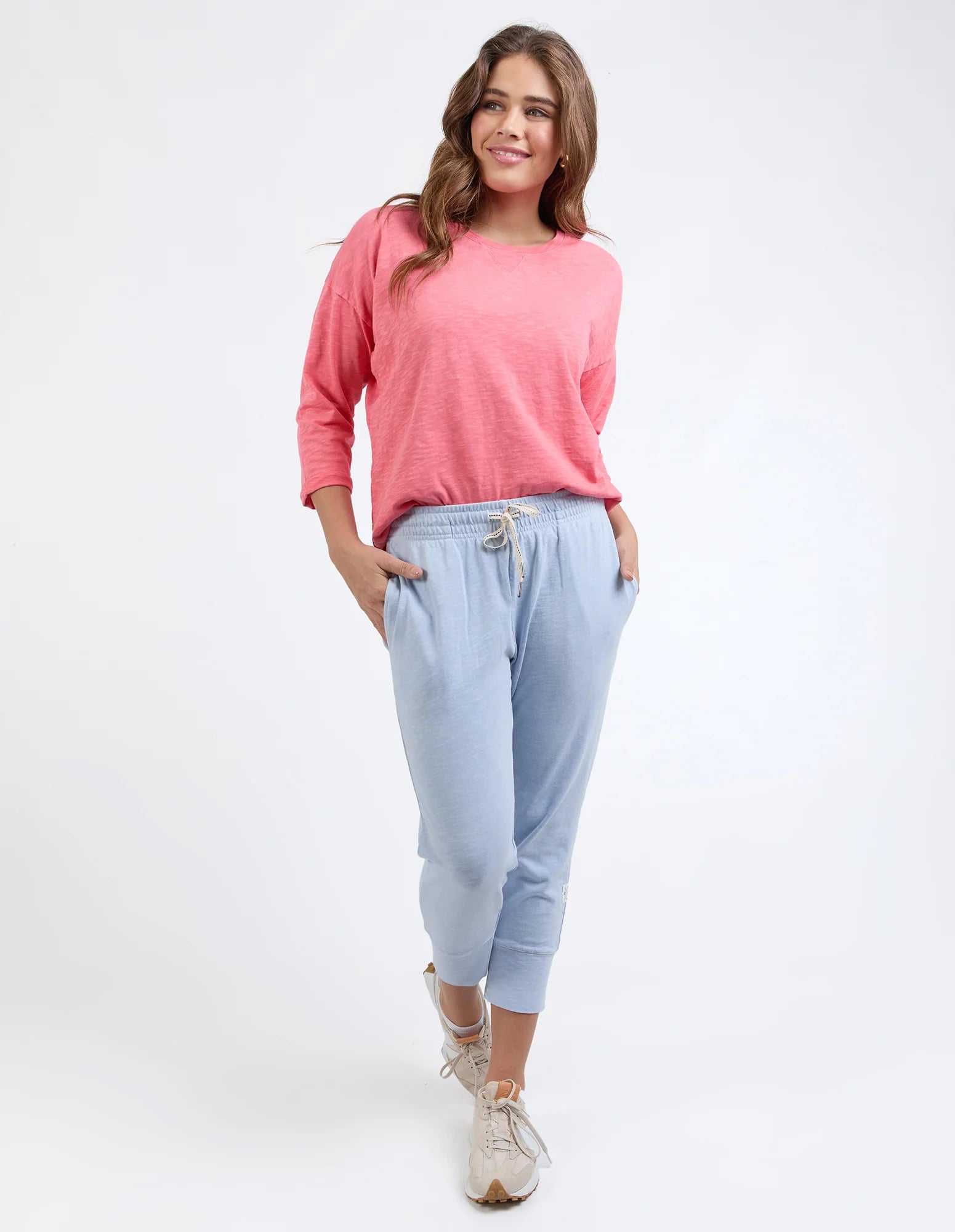 Image of A woman modeling a pink long-sleeve crewneck top and Elm light blue jogger pants with beige sneakers, standing against a plain white studio background.