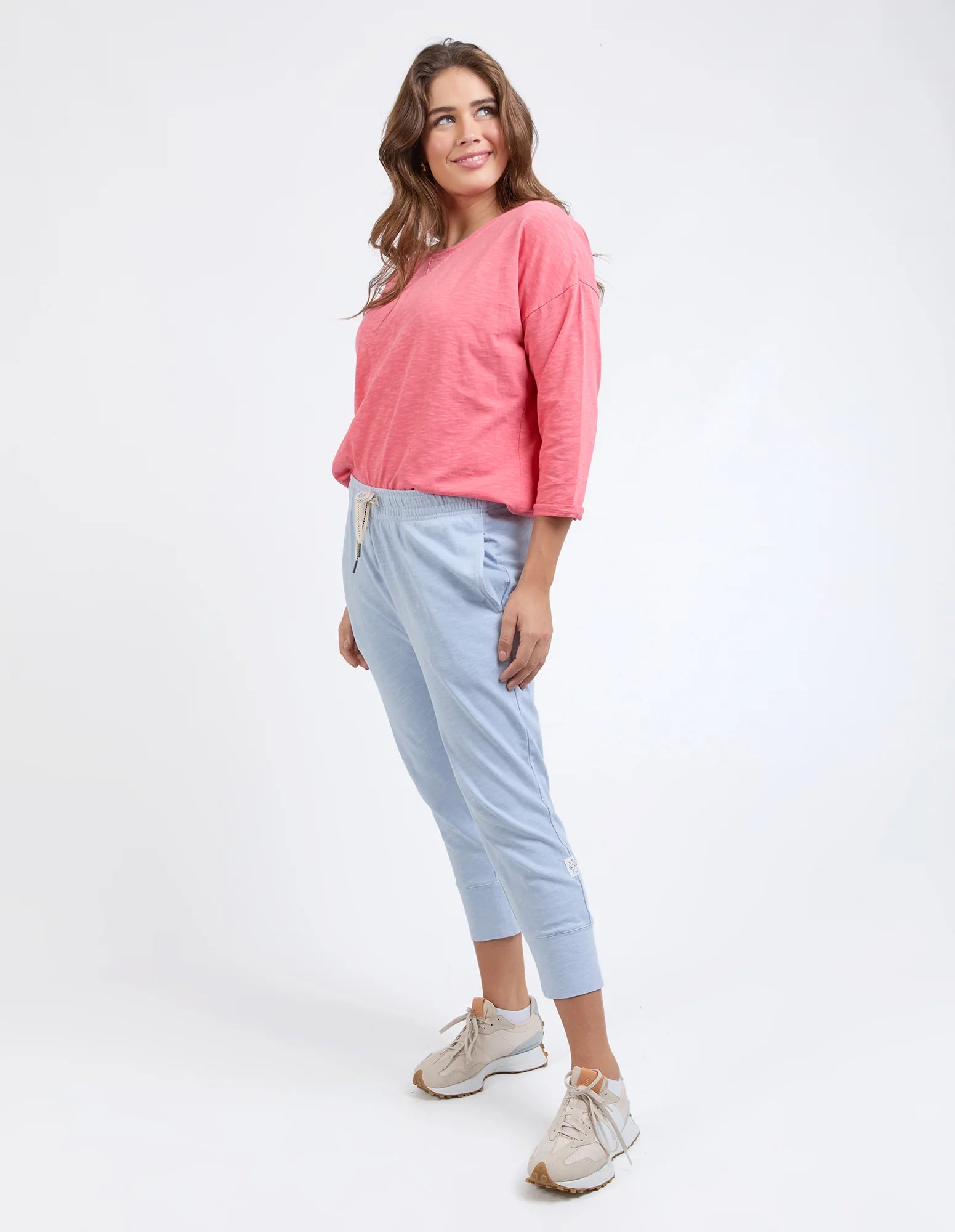 Image of A woman modeling a pink long-sleeve top and Elm light blue cropped pants with beige sneakers, photographed against a plain white studio background.